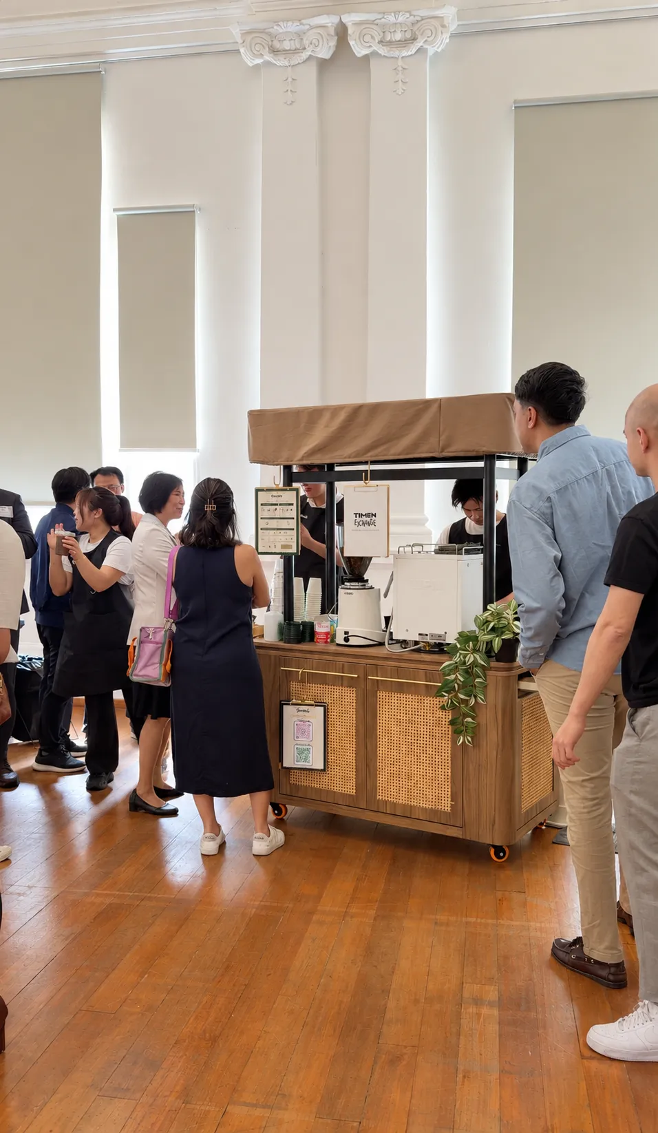 Guests queuing at the Genista Ln kopi cart during a corporate event in a heritage hall, with two baristas serving from behind the cart.