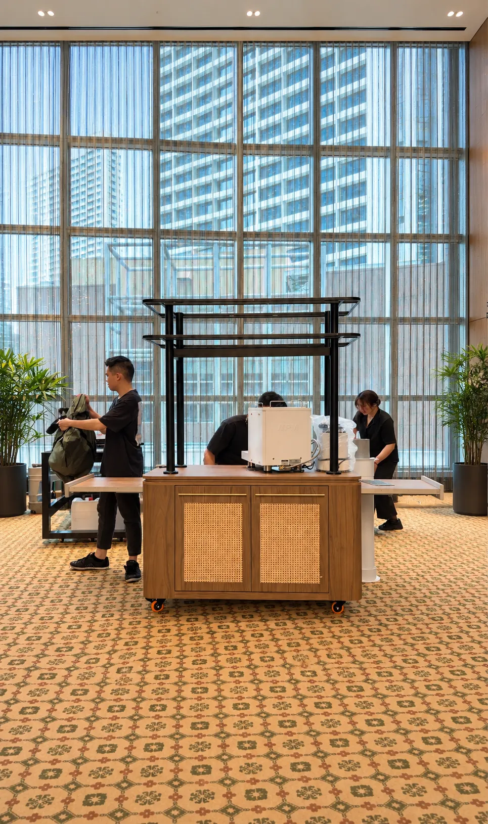 Three Genista Ln crew setting up the Nanyang kopi cart in a Singapore hotel lobby, sliding out the side trays and loading the brew equipment.