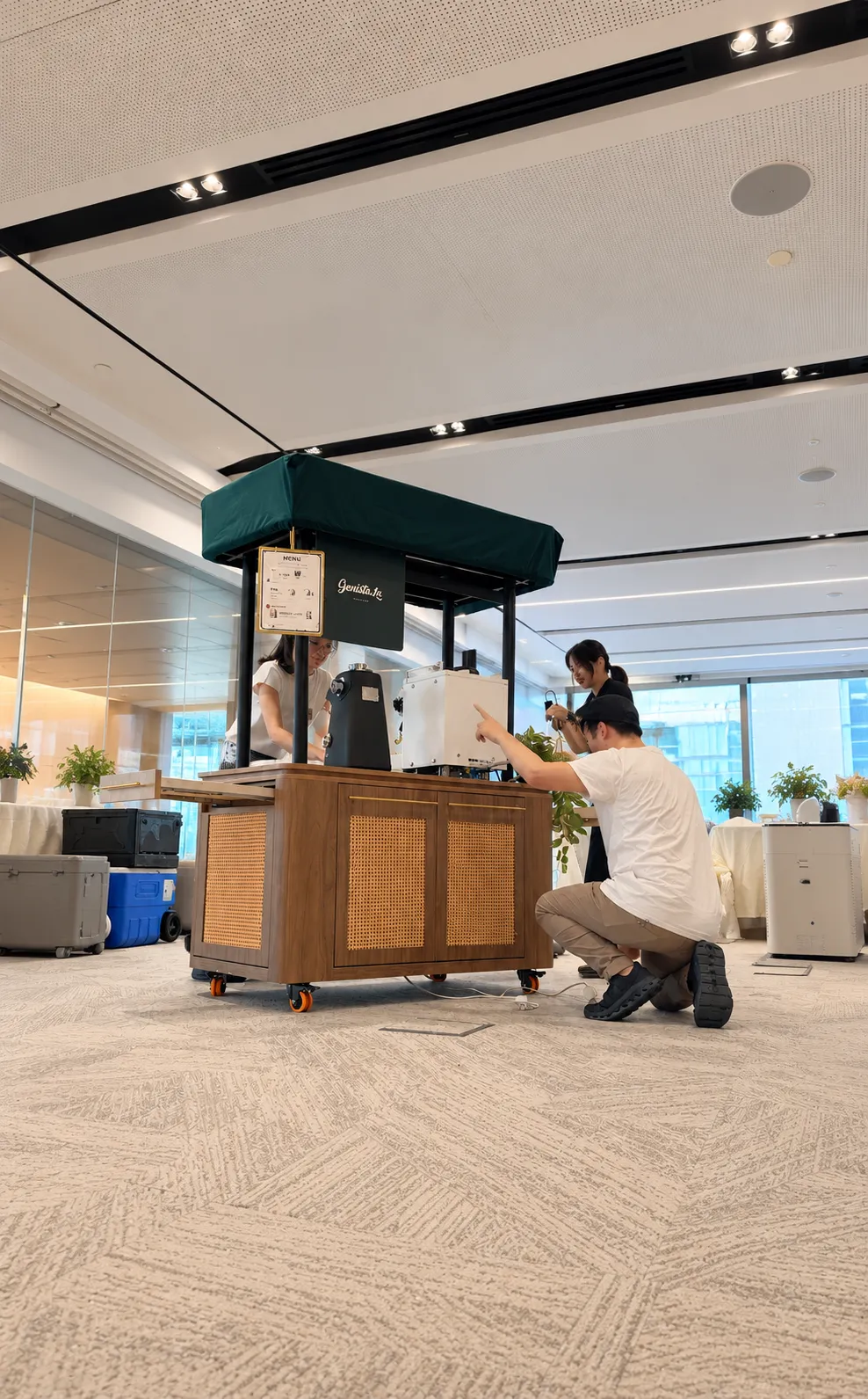 Genista Ln crew assembling the kopi cart under its dark green canopy in an office, one crew member plugging it into a floor power point.
