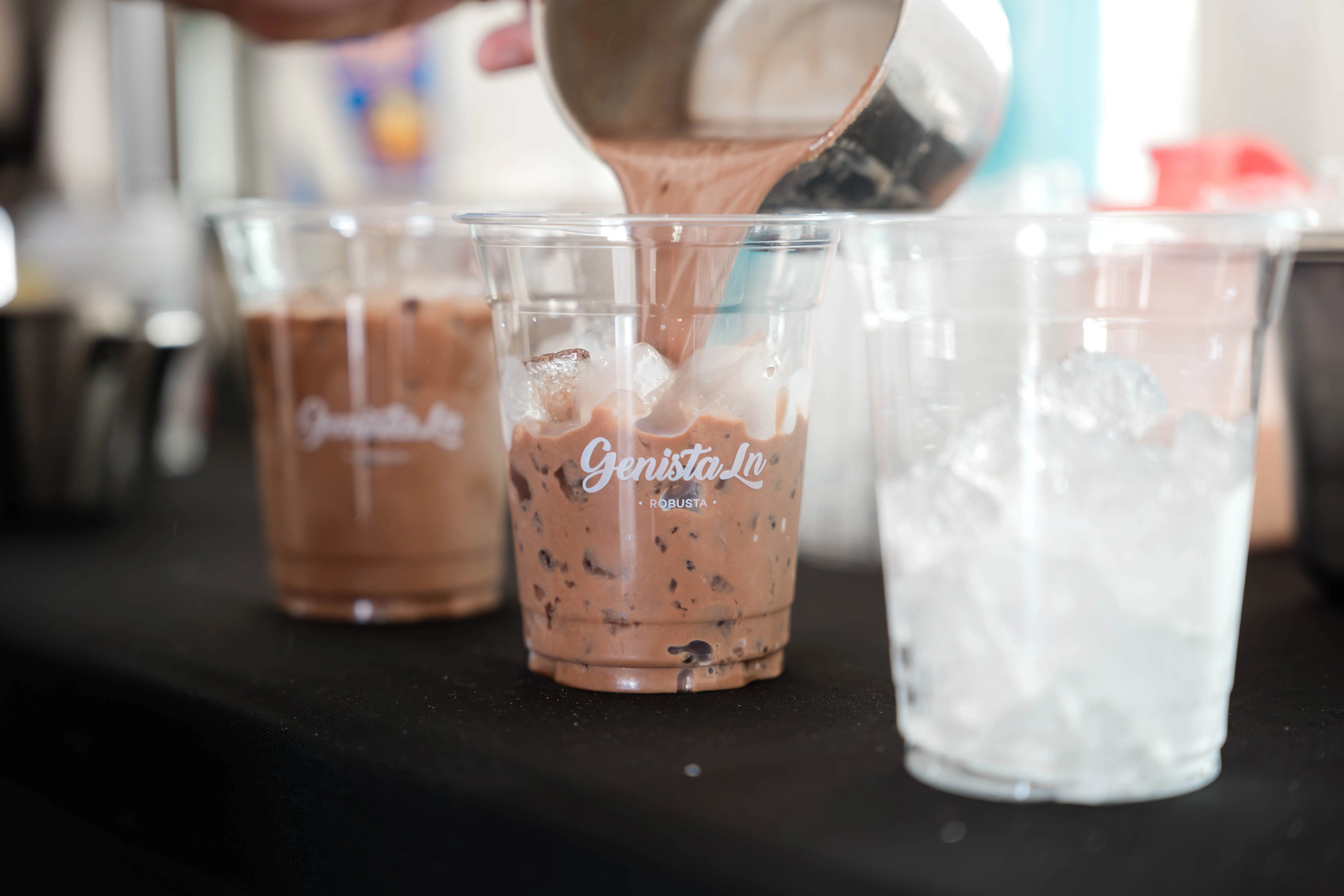 Iced kopi being poured from a jug into a Genista Ln Robusta cup packed with ice, with two more cups on the counter alongside.