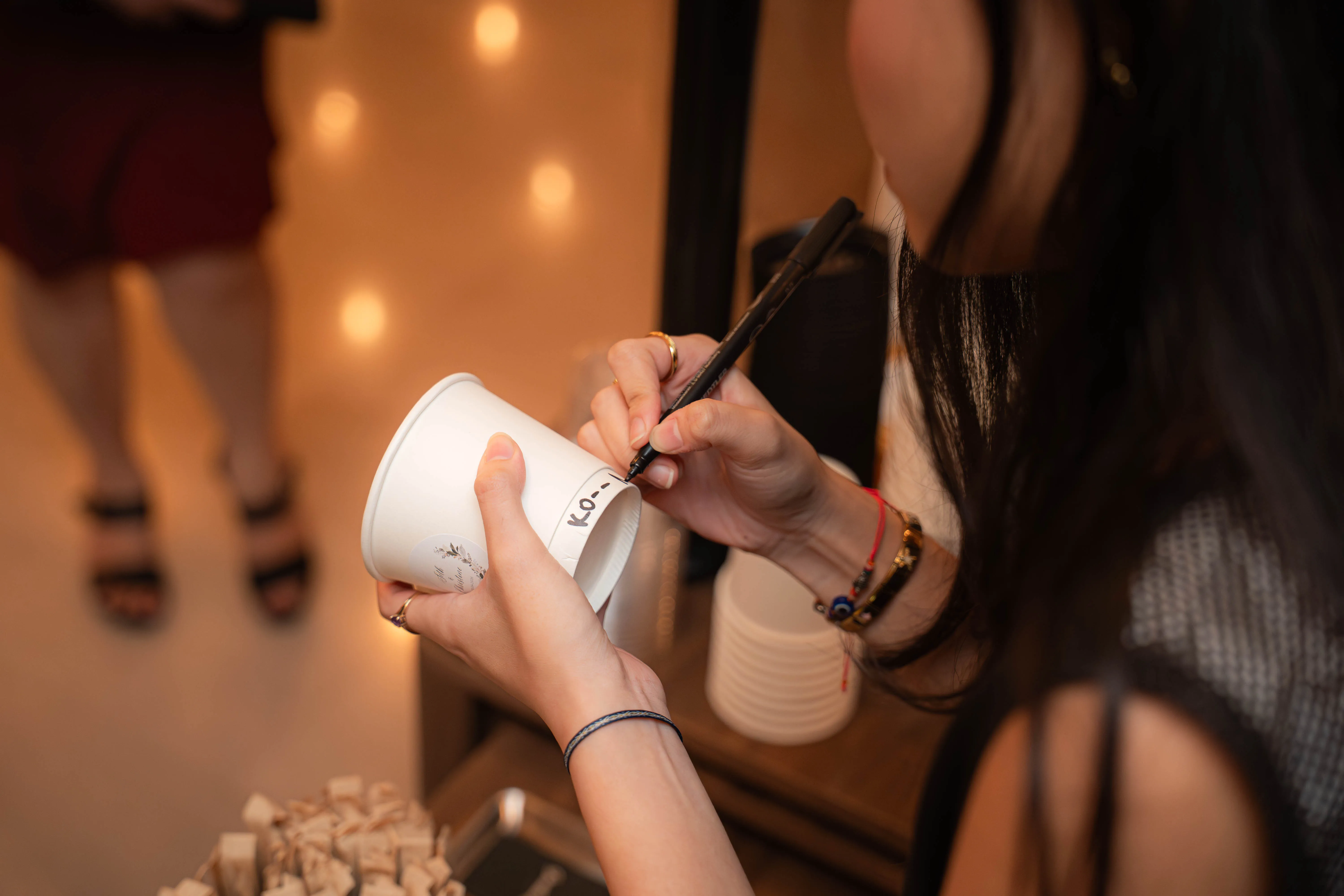 A Genista Ln barista writing a guest’s kopi order onto a paper cup in marker pen at an evening event.