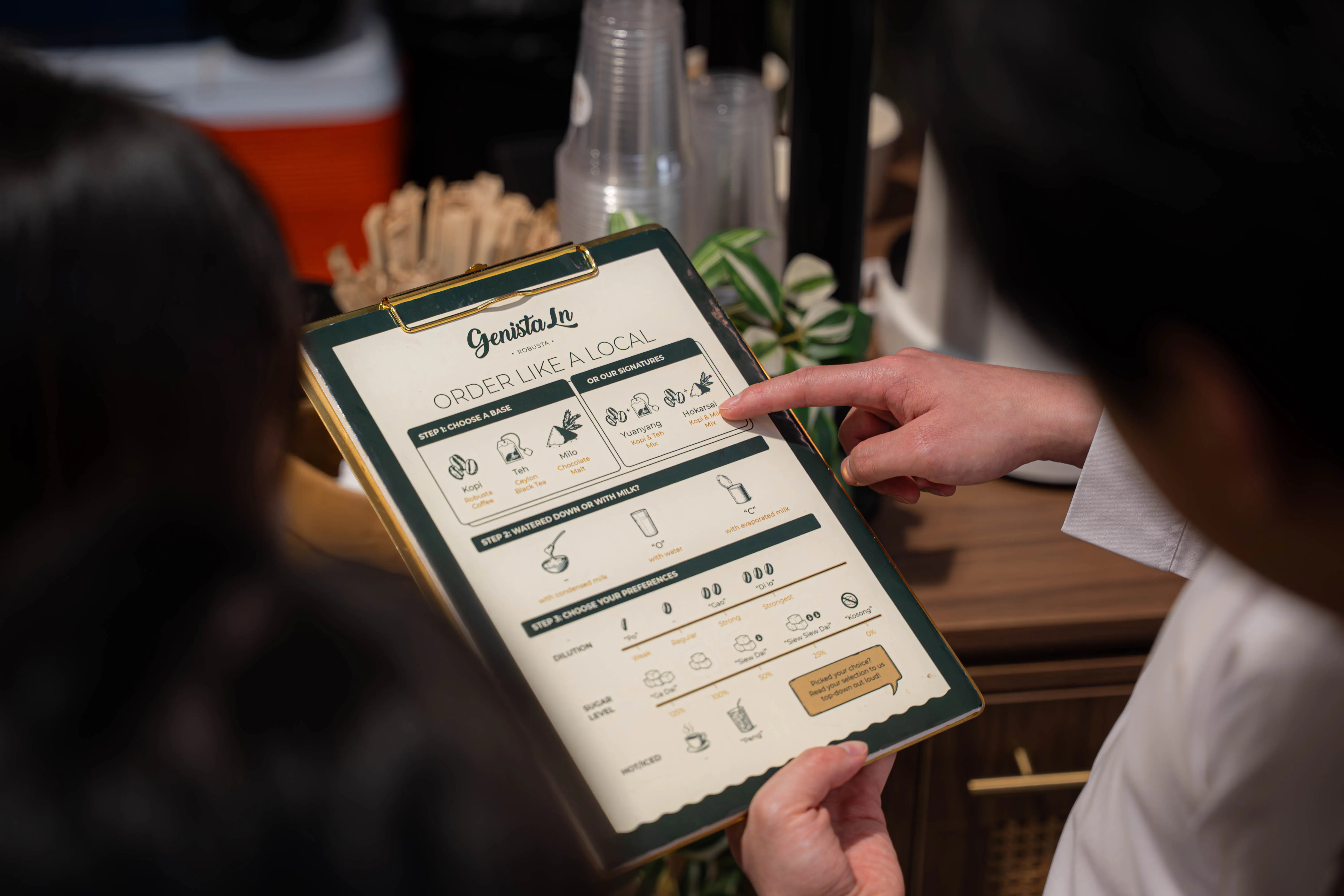 A guest pointing at the Genista Ln “Order Like a Local” card, which walks through choosing a base, then how it is diluted, then the sugar level.