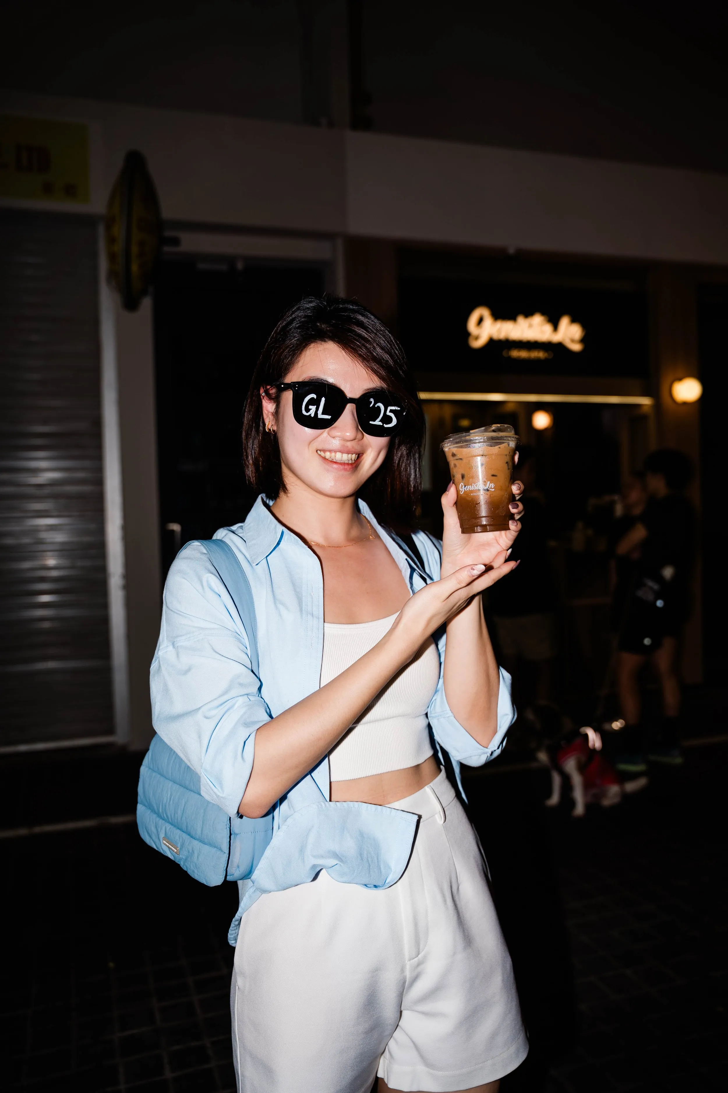 A young woman smiling, wearing sunglasses with 'GL 25' on the lenses, holding a cup of iced coffee in front of a dark background with a sign that reads 'Gourmet Inn' at night