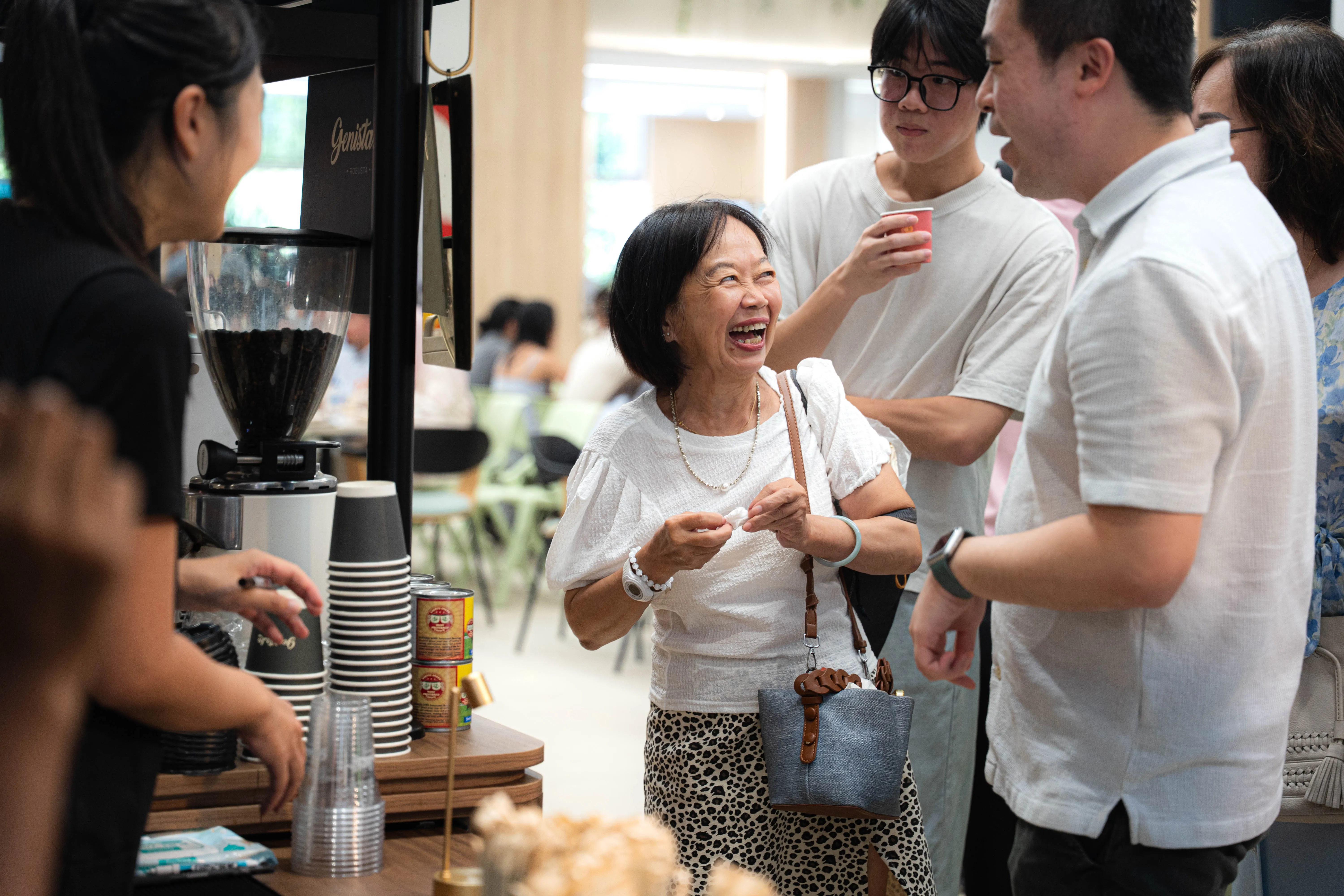 An older woman laughing with the barista across the Genista Ln cart, tins of condensed milk and a stack of cups on the counter between them.