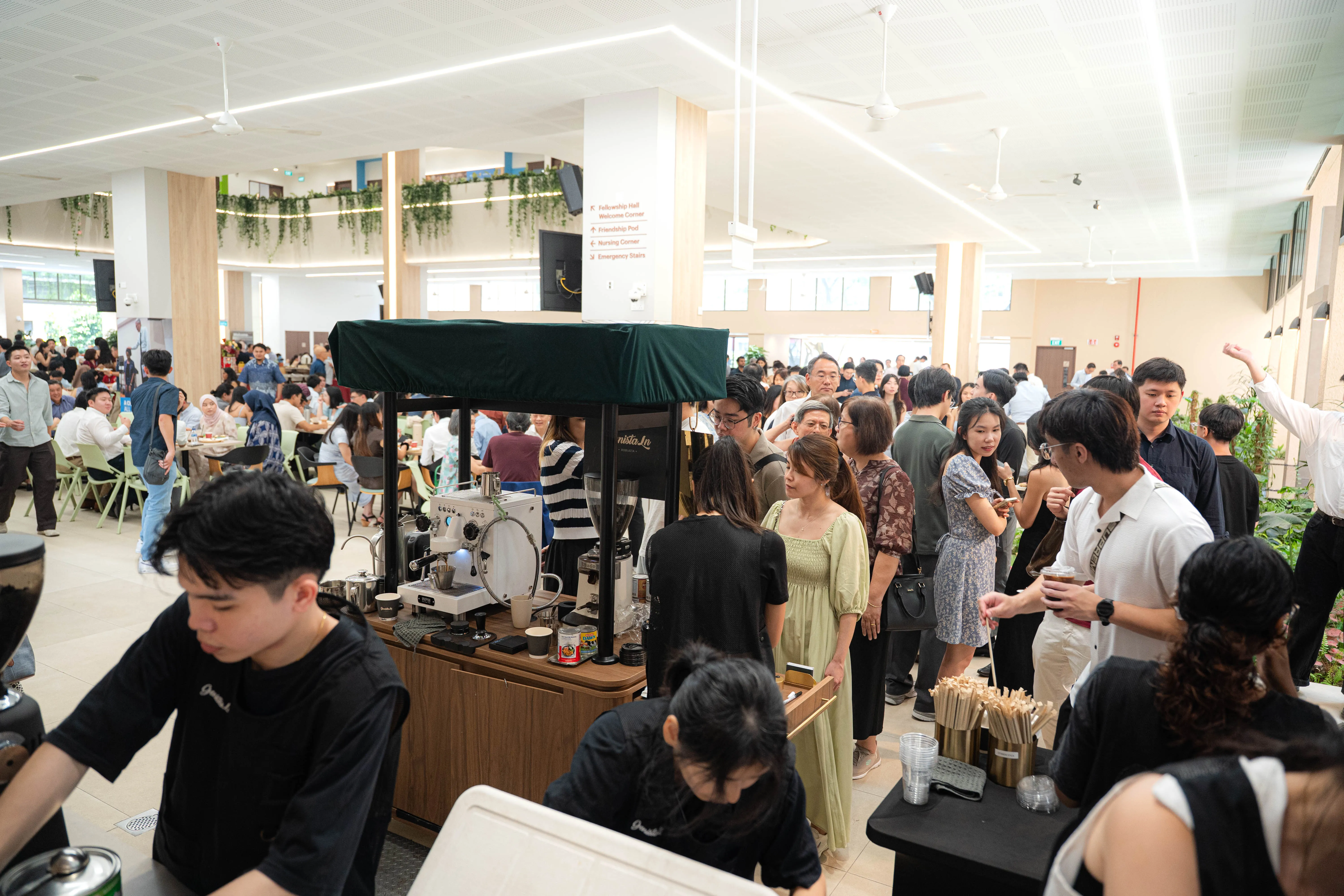 A long queue of guests of every age at the Genista Ln kopi cart in a packed church fellowship hall, with two crew serving behind the counter.