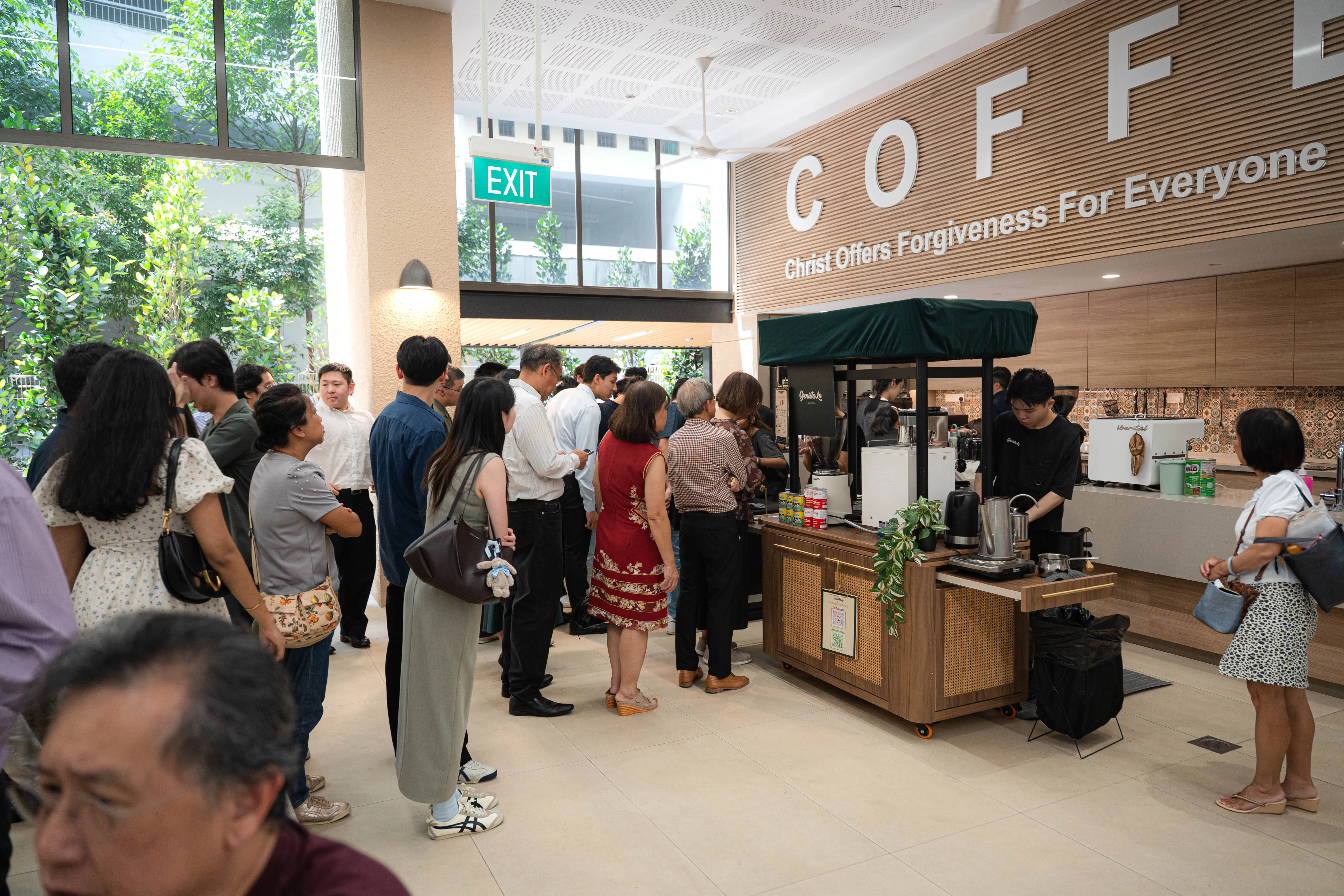 A queue of guests waiting at the Genista Ln kopi cart in a church atrium, below a wall reading “Christ Offers Forgiveness For Everyone”.