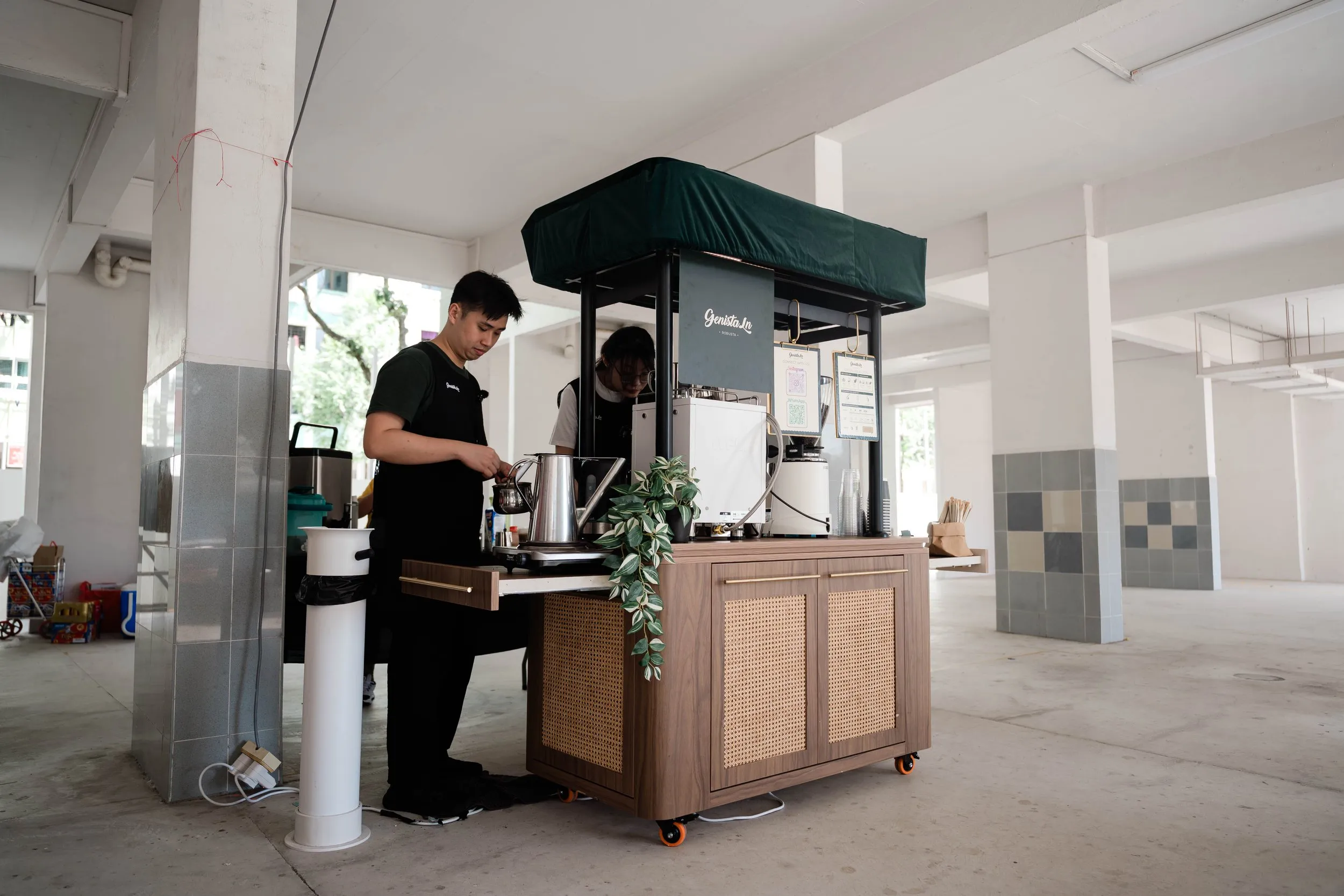 A small coffee stand set up in an open, airy space with two staff members preparing beverages. The stand has a wooden base, a black canopy, and menus hanging on the side