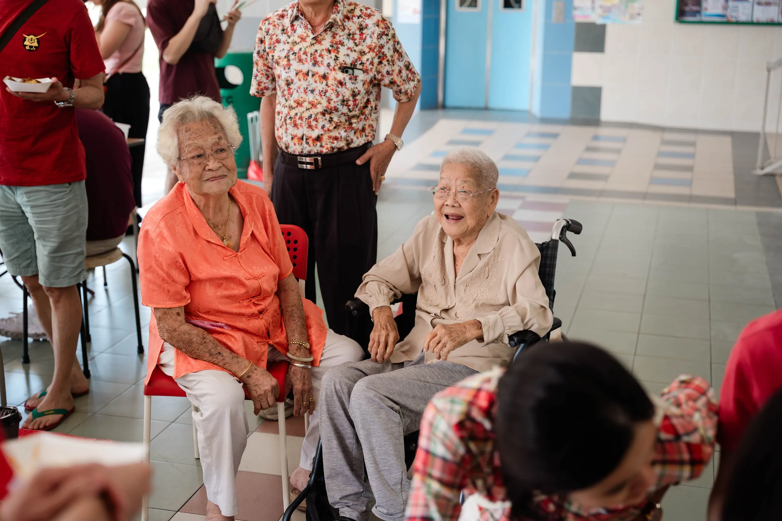 Two elderly women, one sitting on a chair and the other in a wheelchair, are talking to each other and smiling. Several people are standing and walking in the background in a brightly lit indoor setting under a void deck