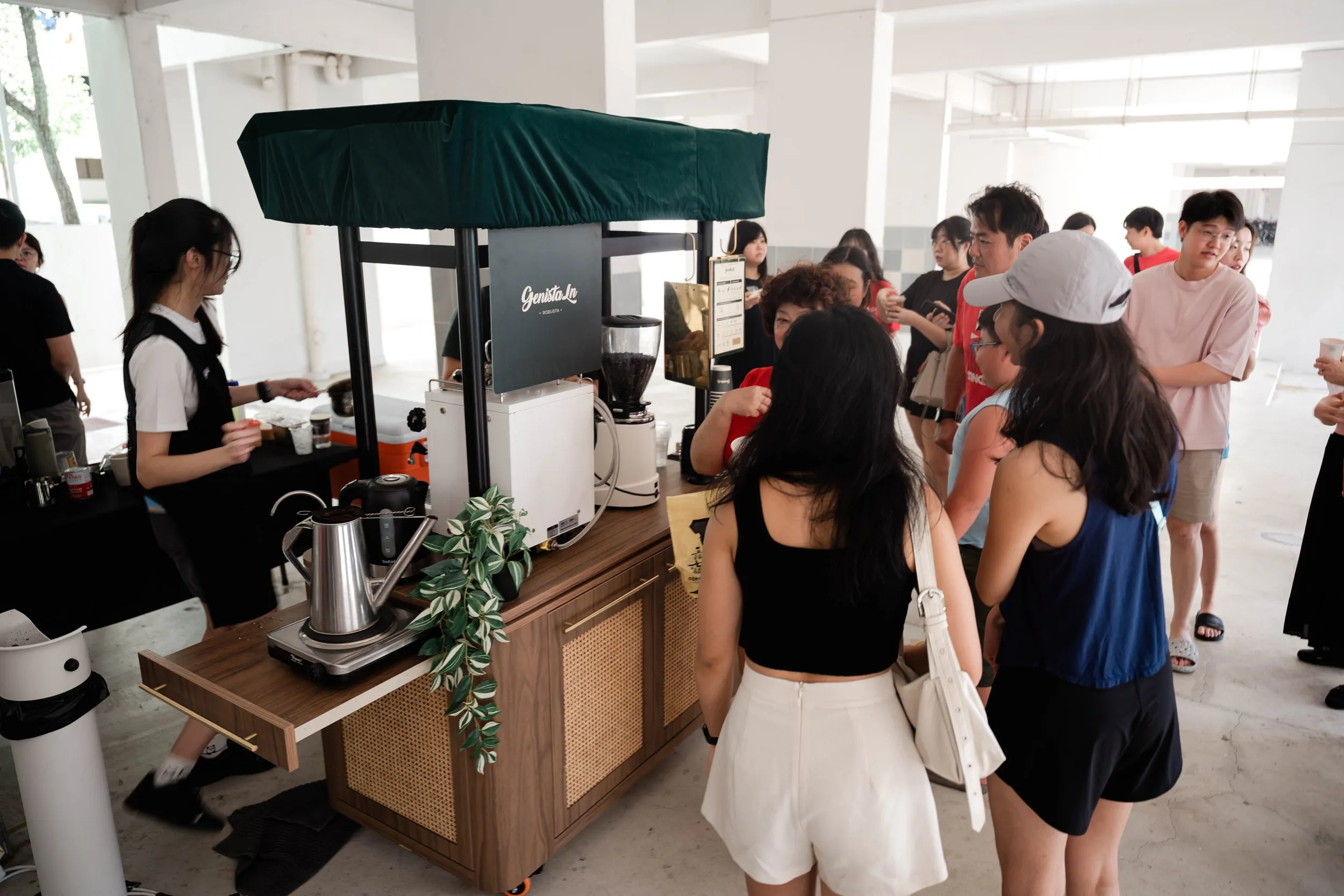 People standing in line at a coffee stand under a void deck, with barista preparing drinks and coffee equipment on the counter