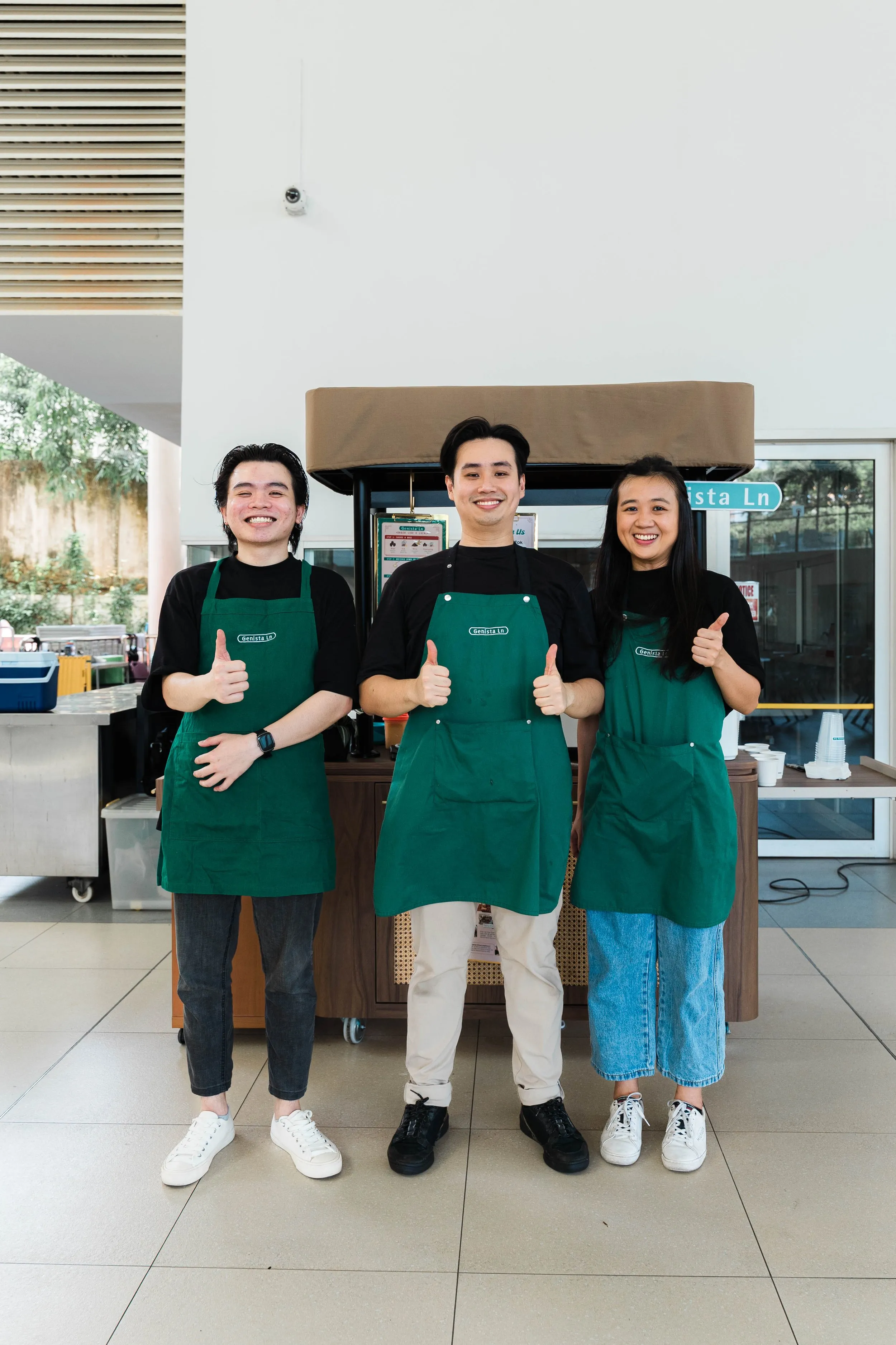 Three smiling employees wearing green aprons standing behind a cafe counter, showing thumbs up