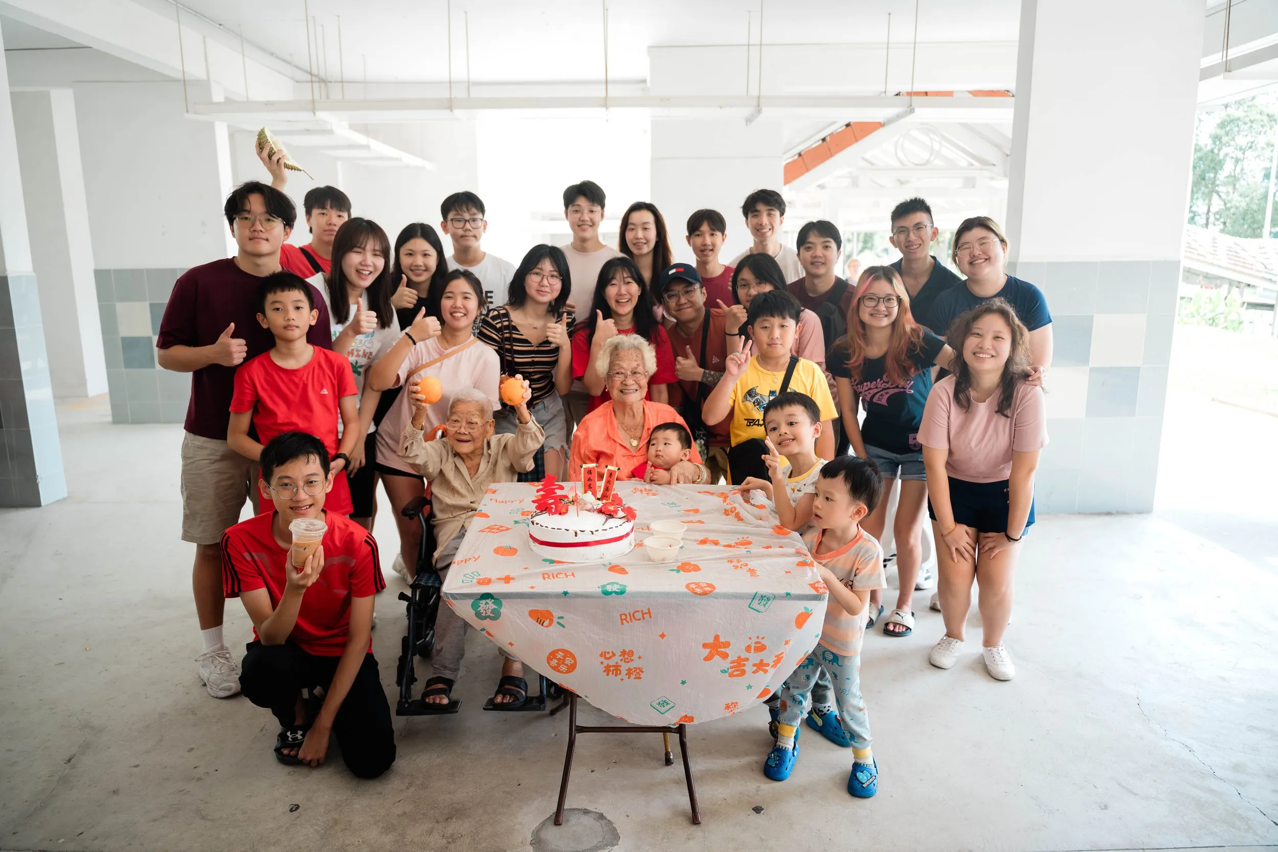 Group of people gathered around a table with a birthday cake, celebrating a birthday with elderly women, children, and adults in a bright, open space