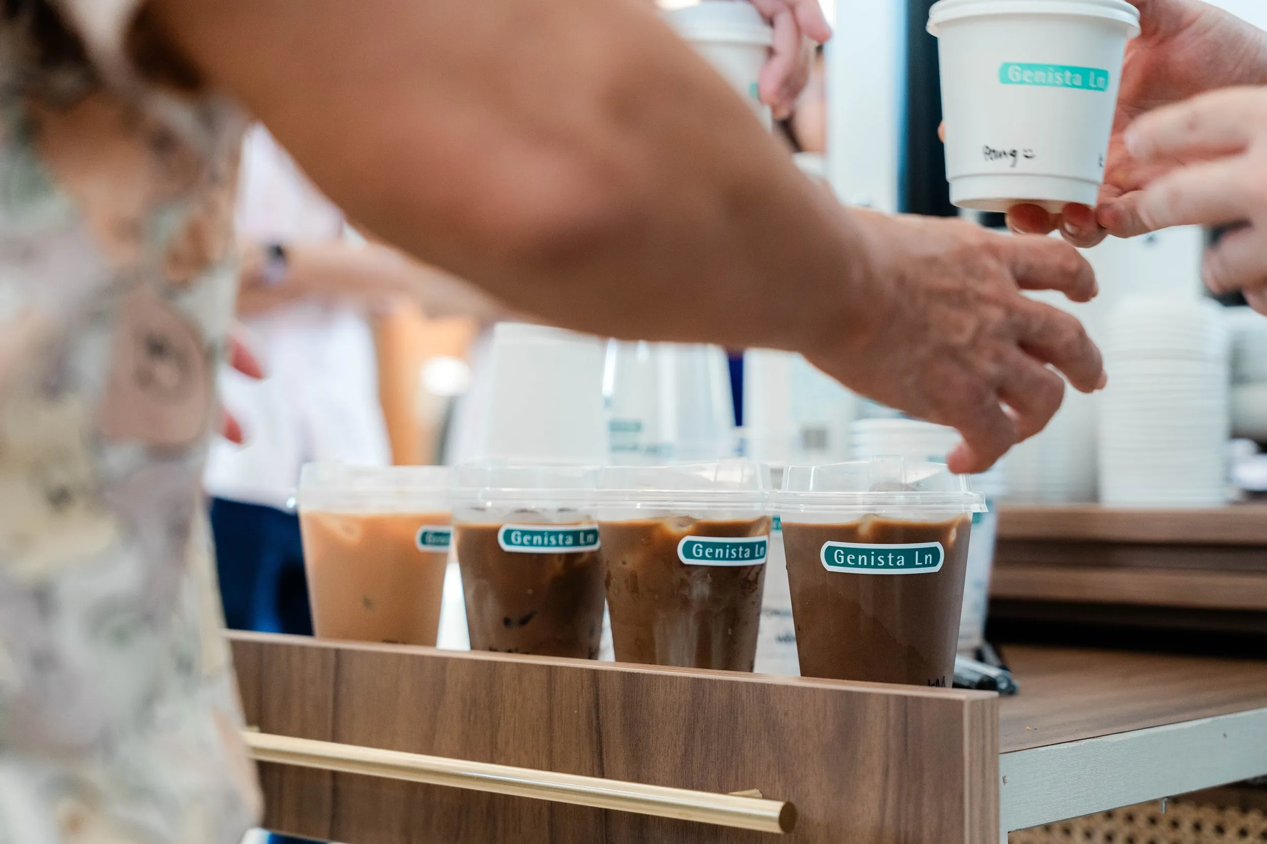 People serving iced coffee drinks in plastic cups with labeled stickers that say "Genista Ln", on a wooden tray at a coffee stand
