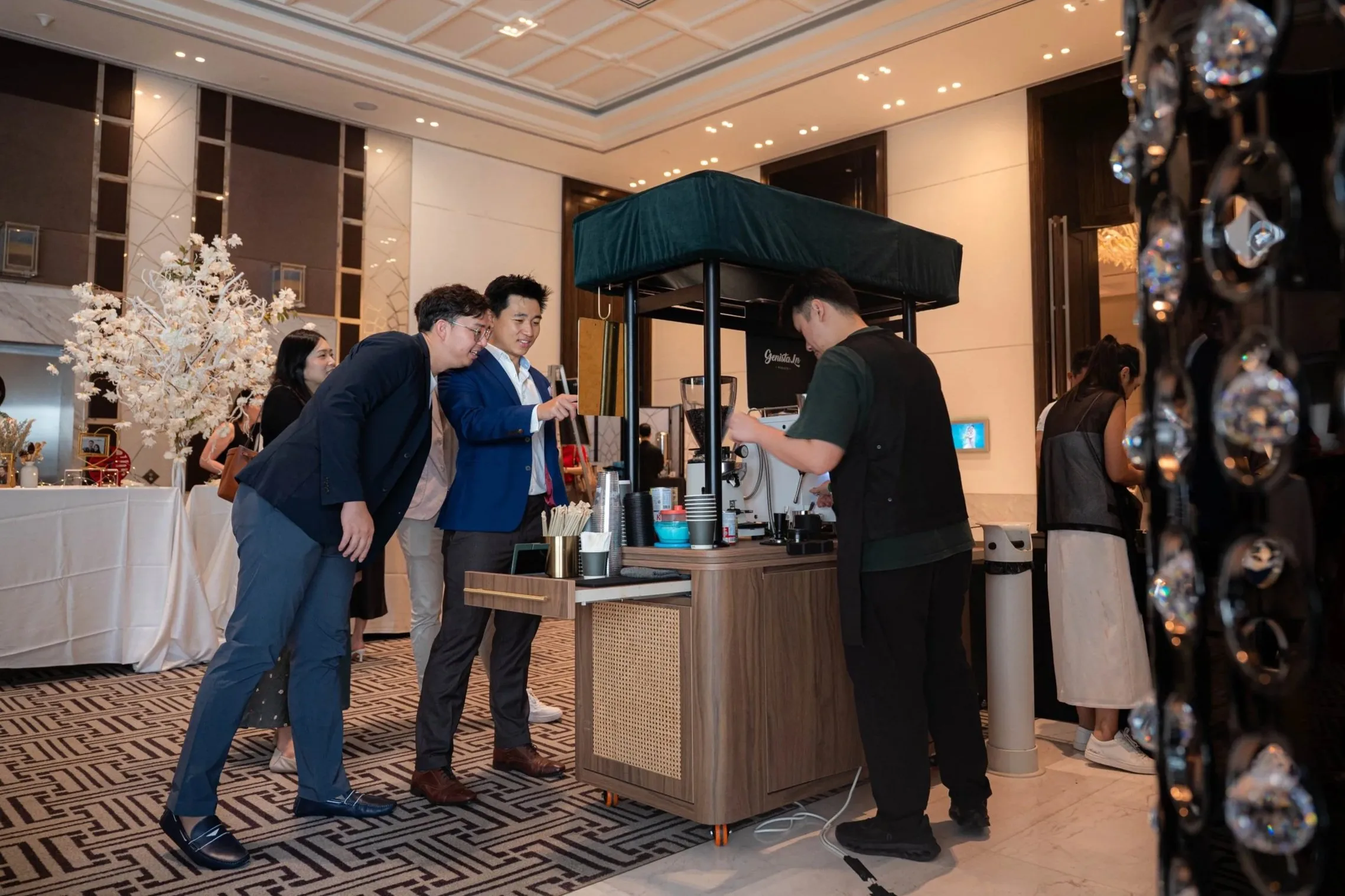 Group of people ordering coffee at Genista Ln Kopi Cart in a hotel lobby in Westin, with ornate decor and floral arrangements visible in the background