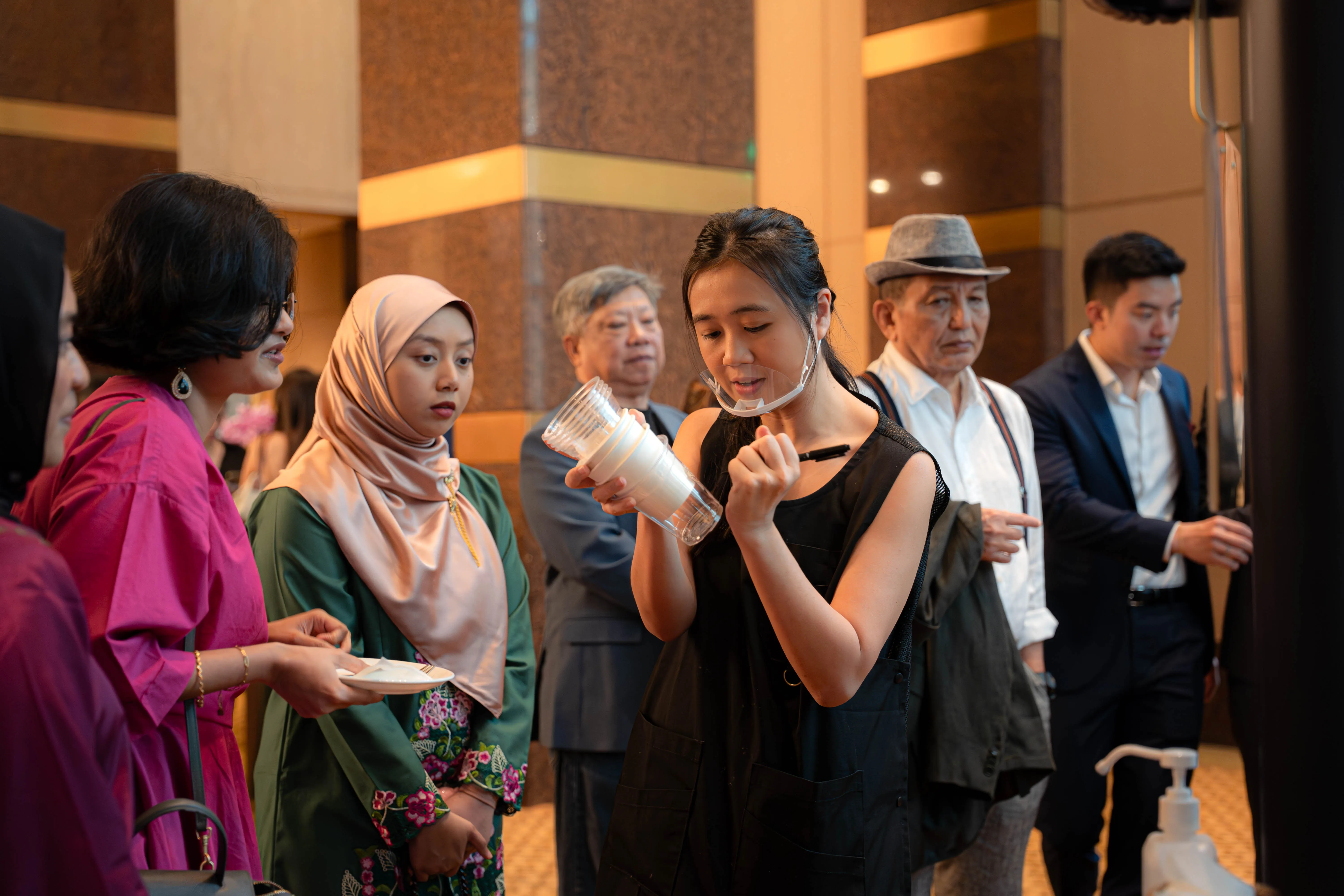 A queue of guests of mixed ages waiting at the Genista Ln cart in a hotel foyer, among them a young woman in a peach hijab and green kebaya and two older men, one in a dark suit and one in a grey fedora.