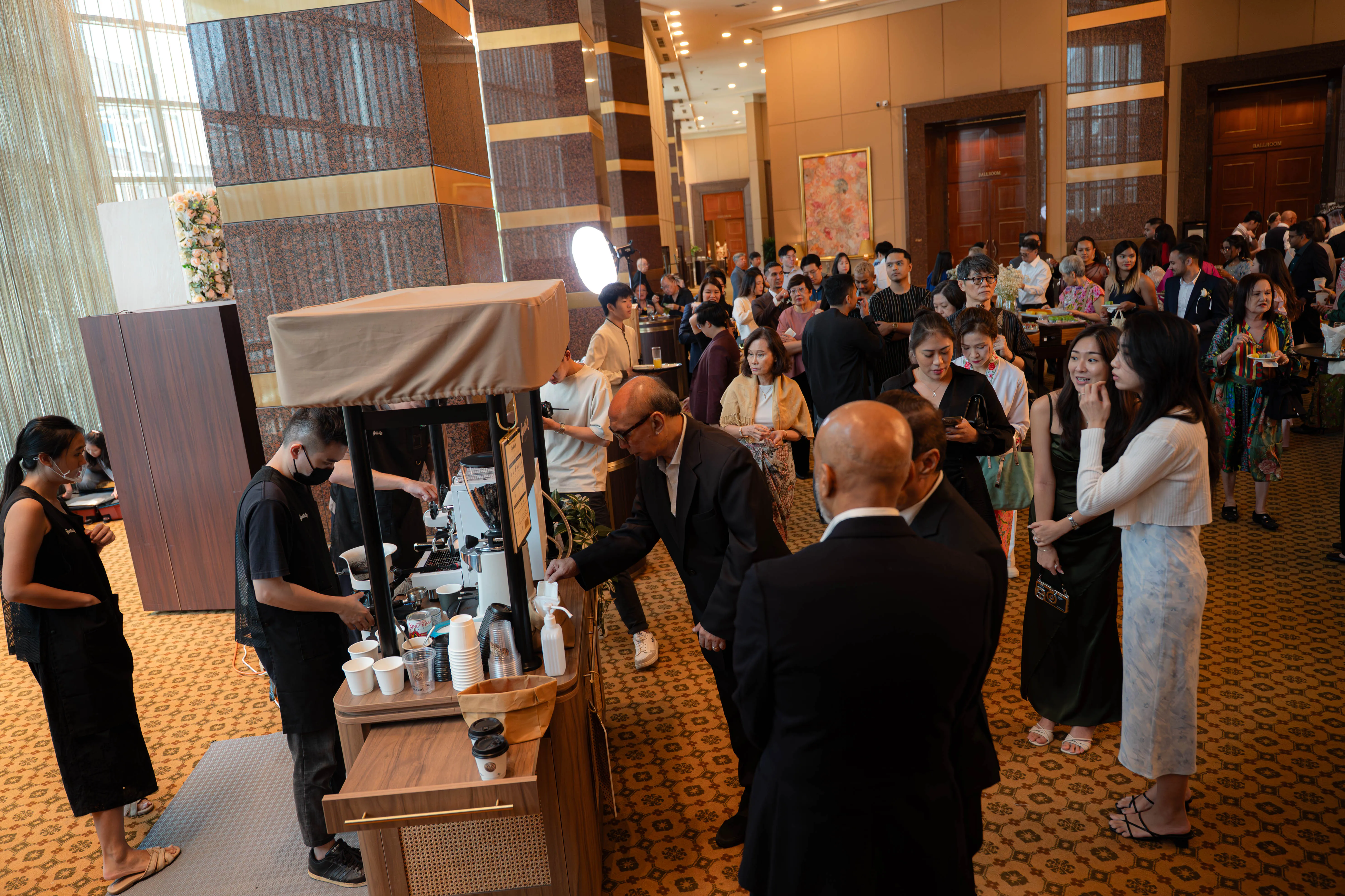Guests crowded around the Genista Ln kopi cart in a hotel foyer outside the ballroom doors, many of them older relatives waiting with cups in hand.
