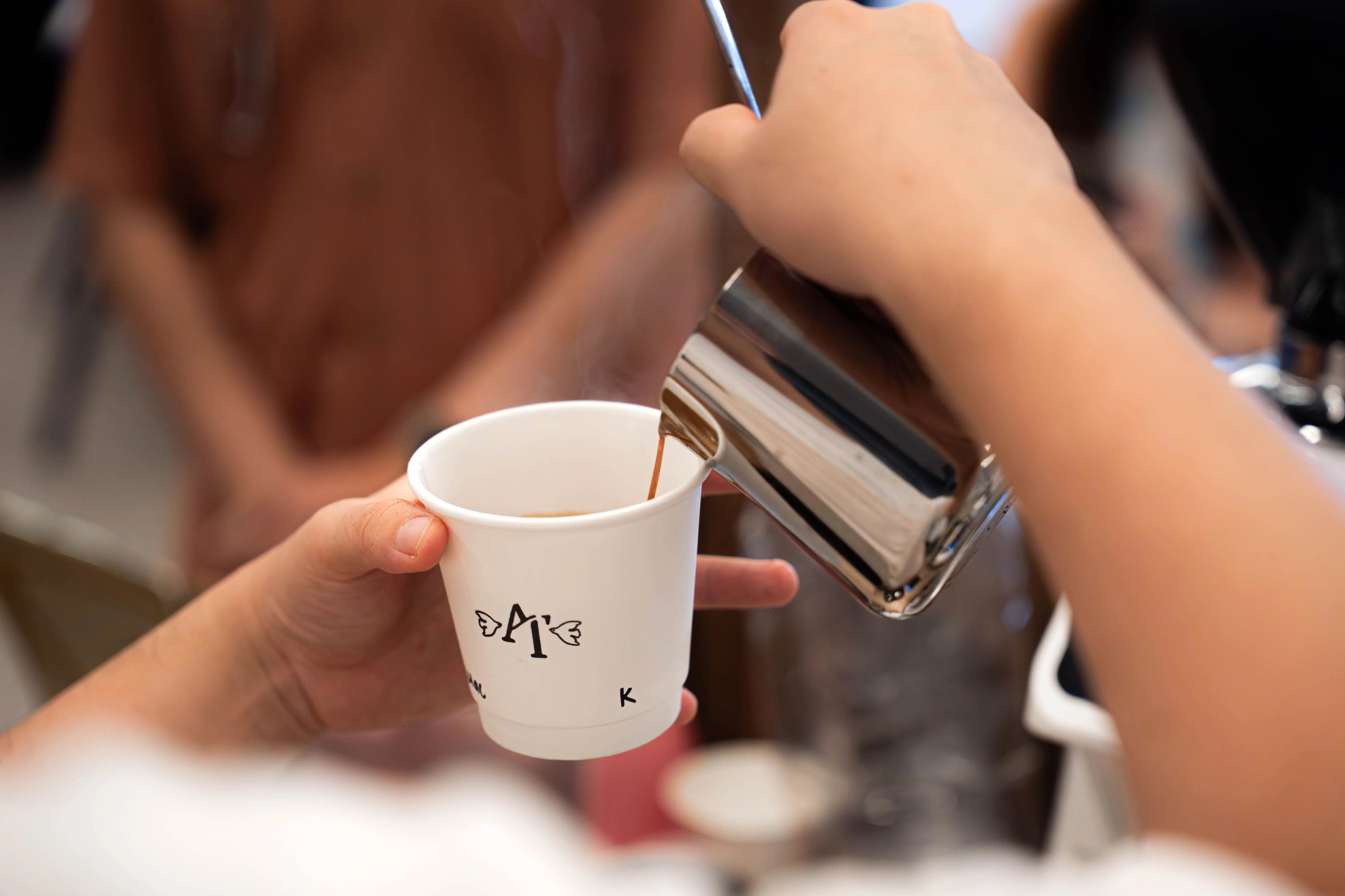 Hot kopi being poured from a metal jug into a white cup carrying the couple’s hand-drawn winged monogram, with a guest’s name written beneath it in marker.