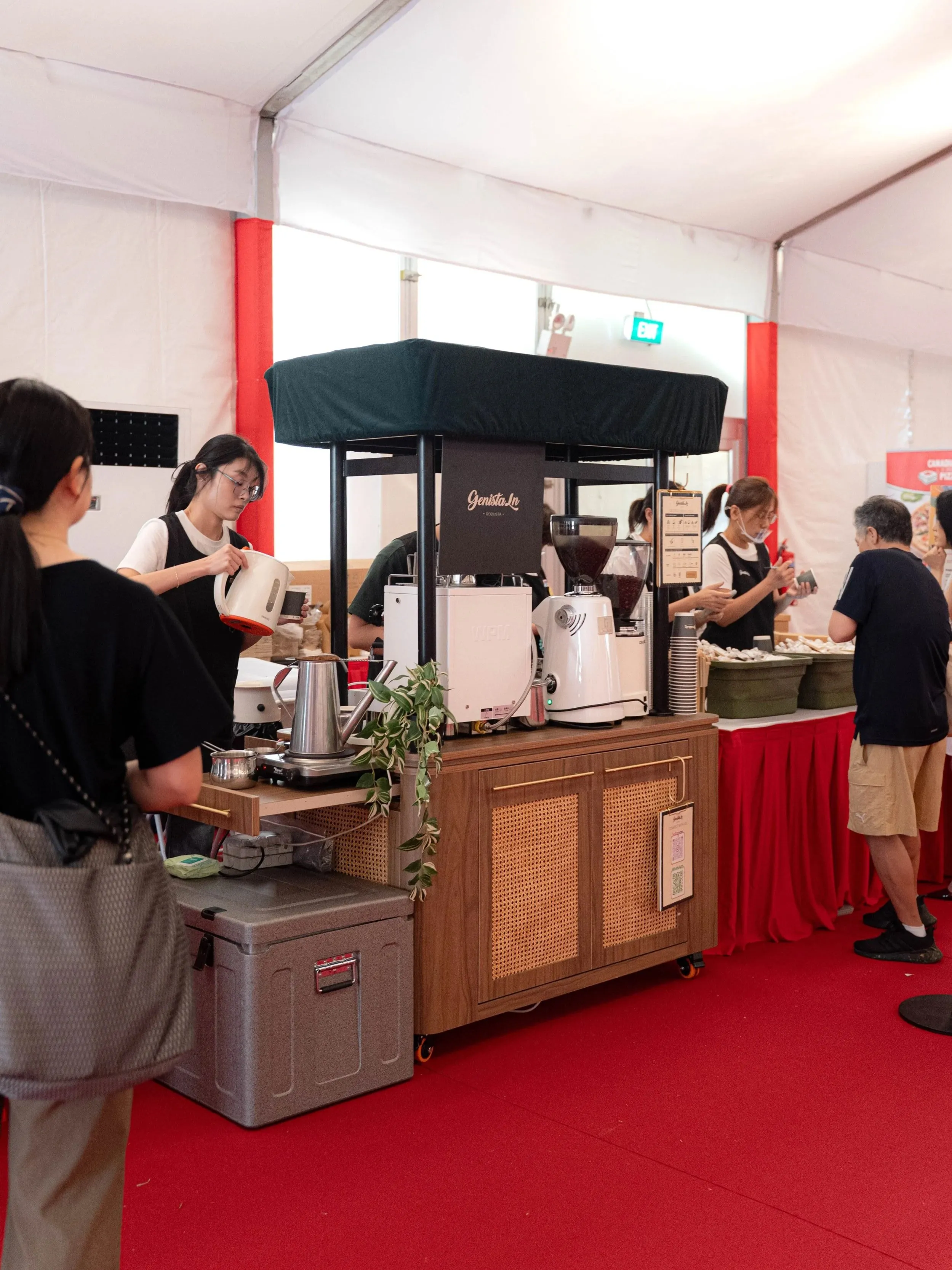 The Genista Ln kopi cart in a large white marquee at The Istana Open House, a crew member pouring from a kettle while visitors queue past red-skirted food tables.