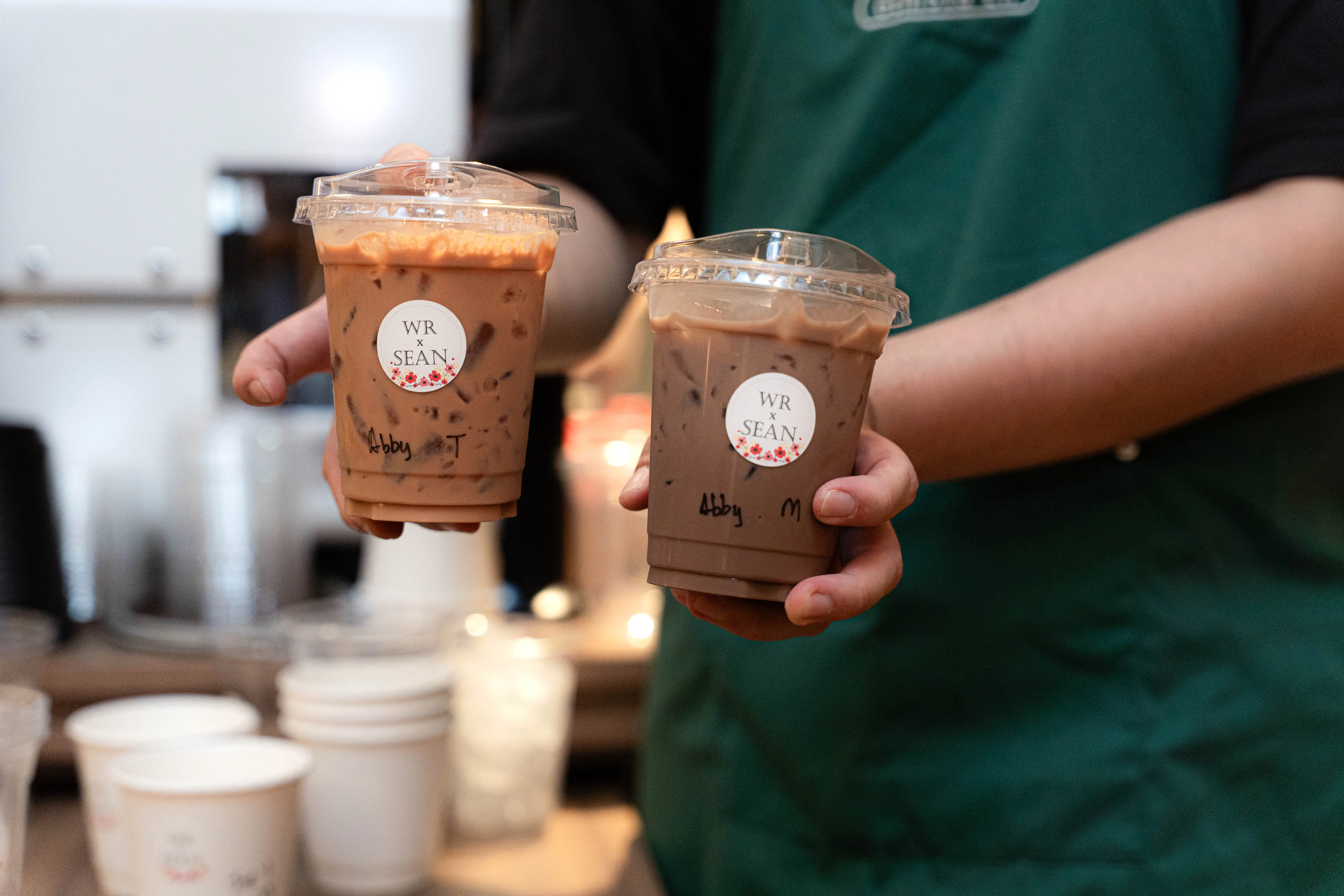 A crew member holding out two iced kopi, each with a floral “WR x SEAN” sticker and a guest’s name written underneath.