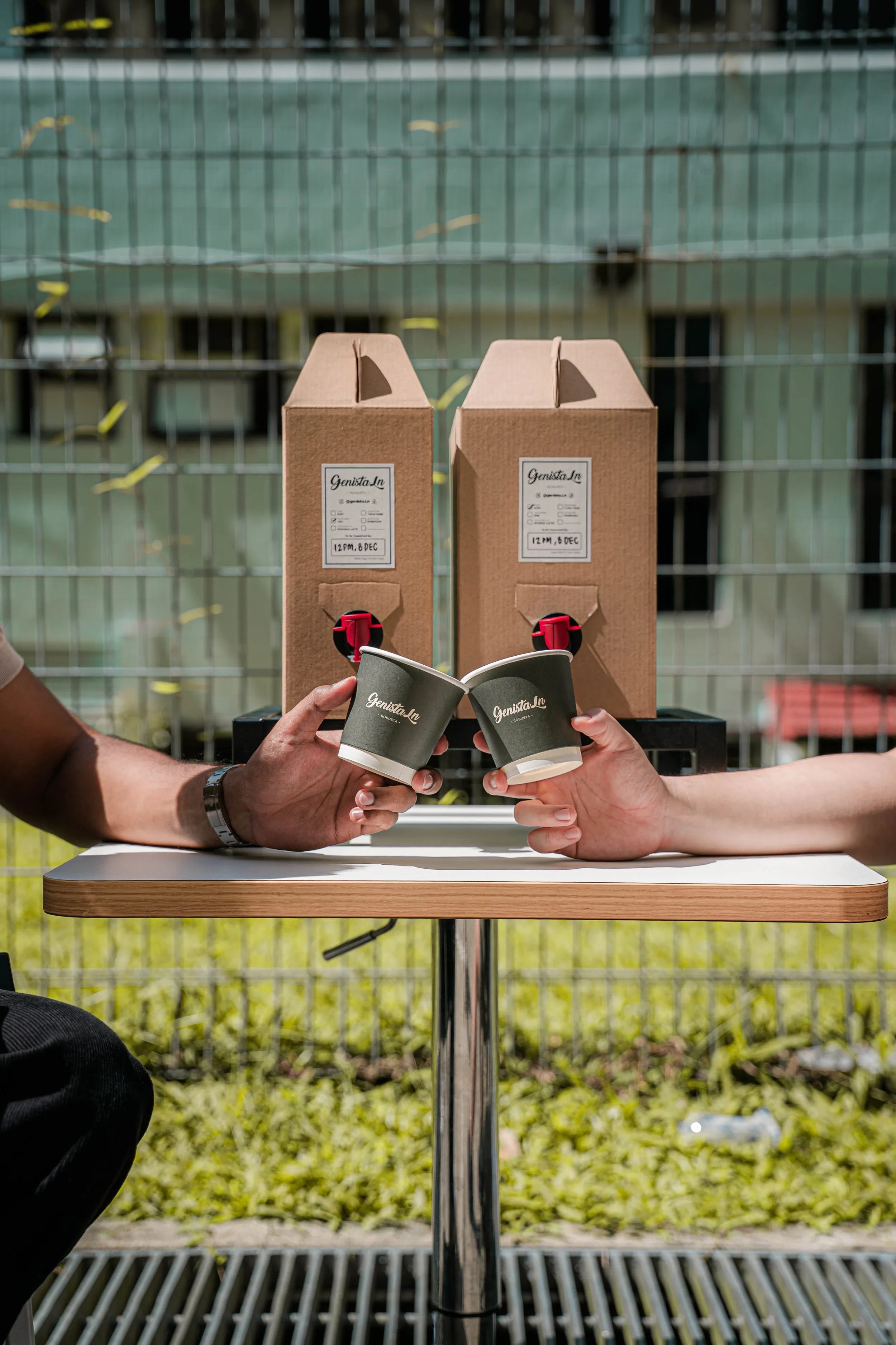 Two Genista Ln party boxes with red taps on an outdoor table, their labels ticked and hand-dated, as two people touch cups together in front of them.