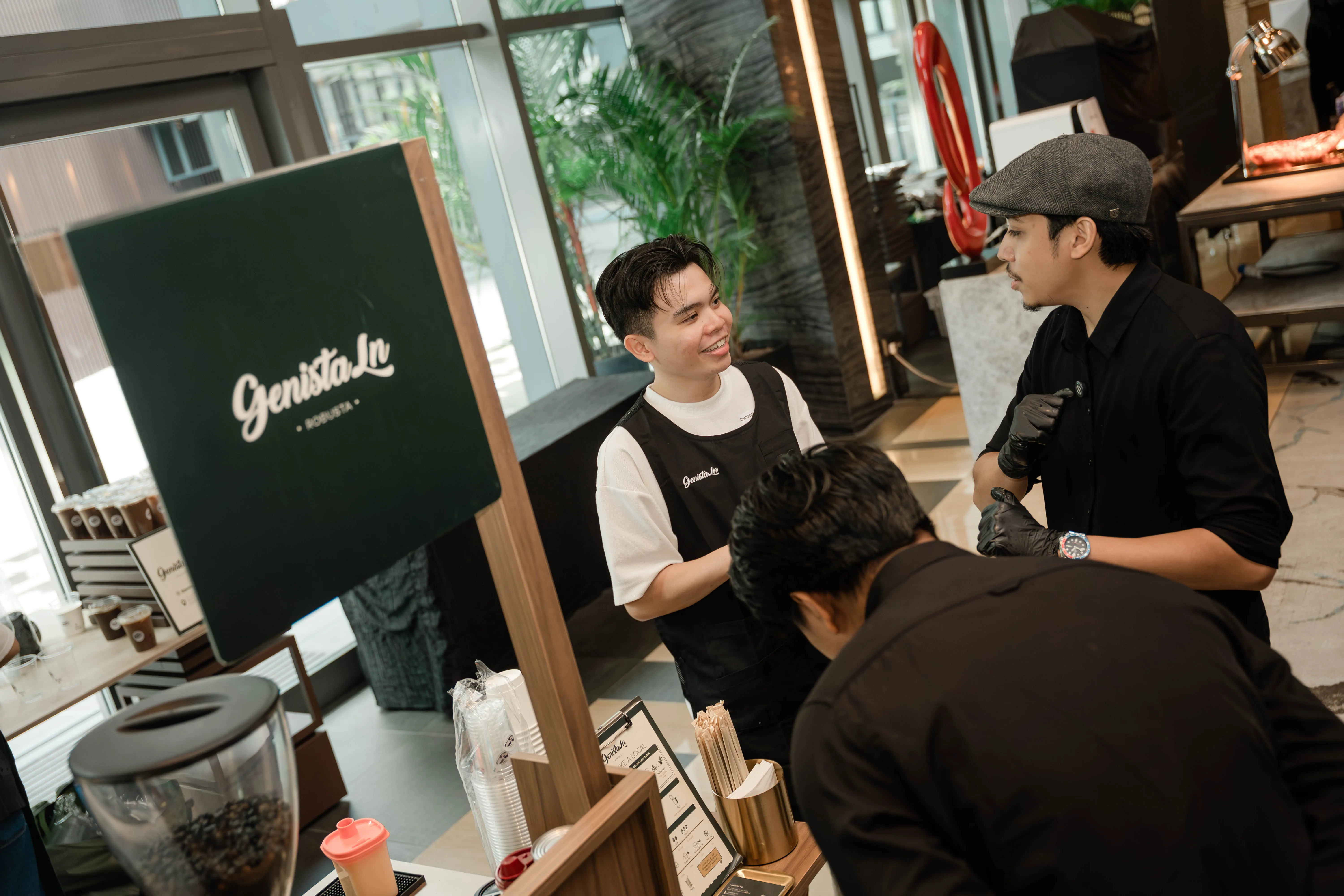 A Genista Ln barista talking through the running order with two venue staff beside the cart, the ordering guide and cup stack laid out on the counter.