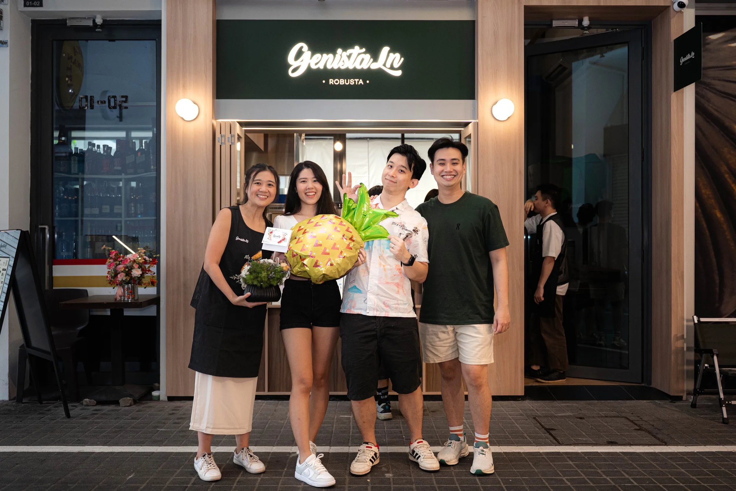 Group of four young adults standing outside a restaurant entrance, celebrating with flowers and a gift, smiling and waving at the camera