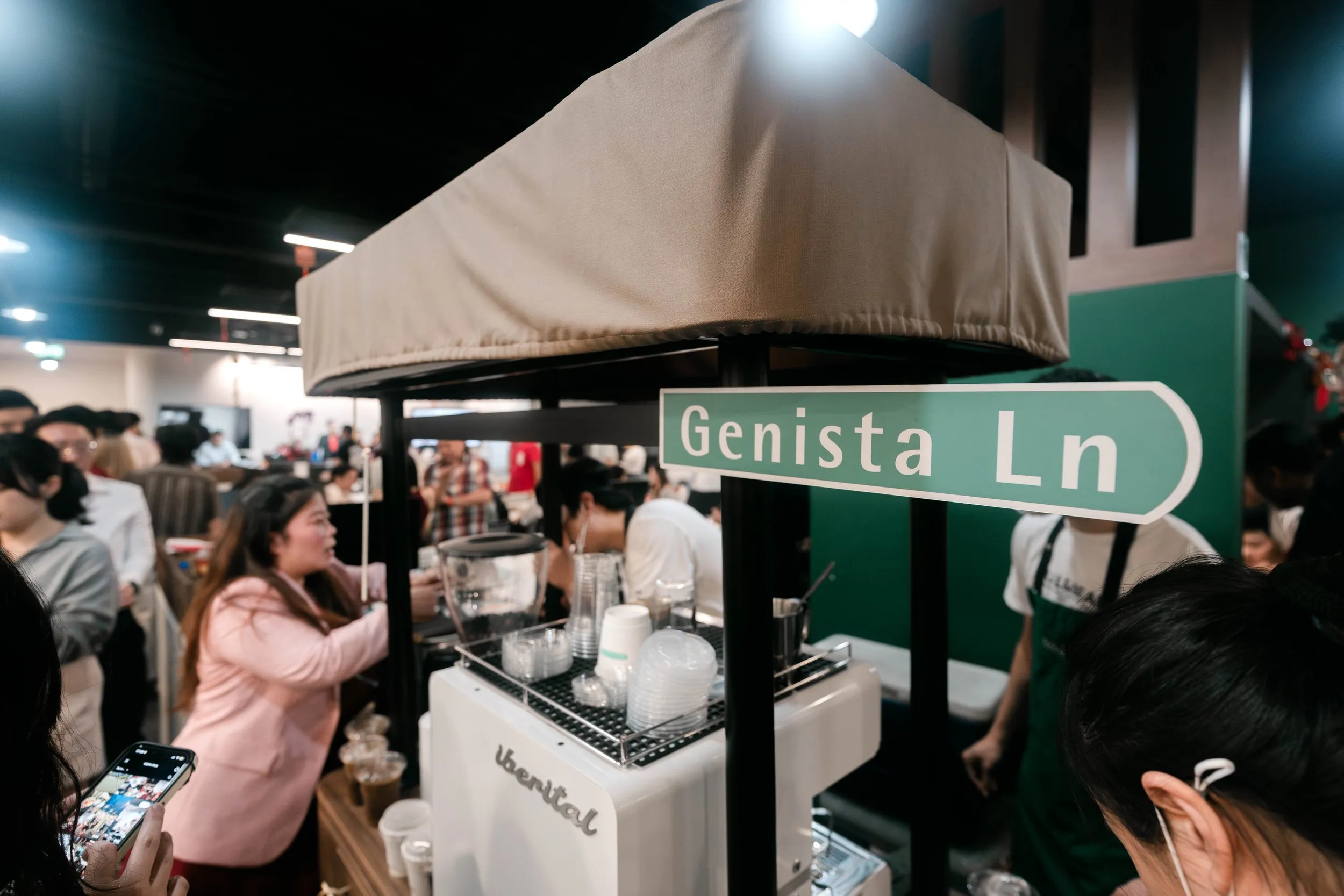 Crowd of people at a Kopi Cart with a sign reading 'Genista Ln' in an indoor setting