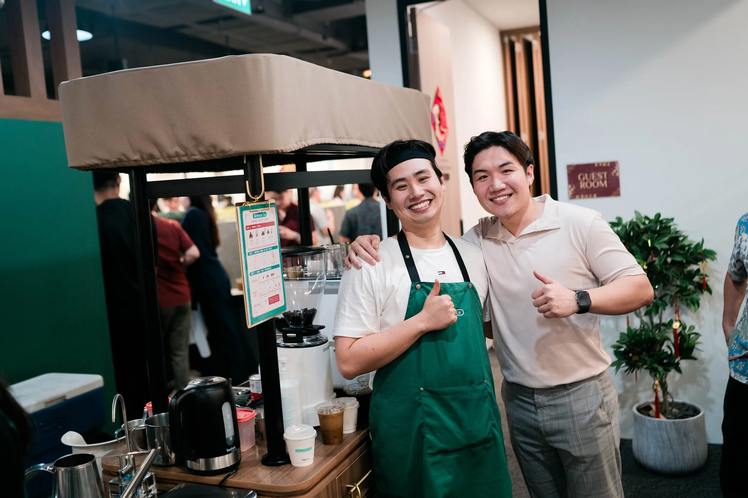 Two men smiling and giving thumbs-up while standing inside a coffee shop. One wears a green apron and black headband, the other a beige polo shirt. Behind them are coffee-making equipment and a potted plant