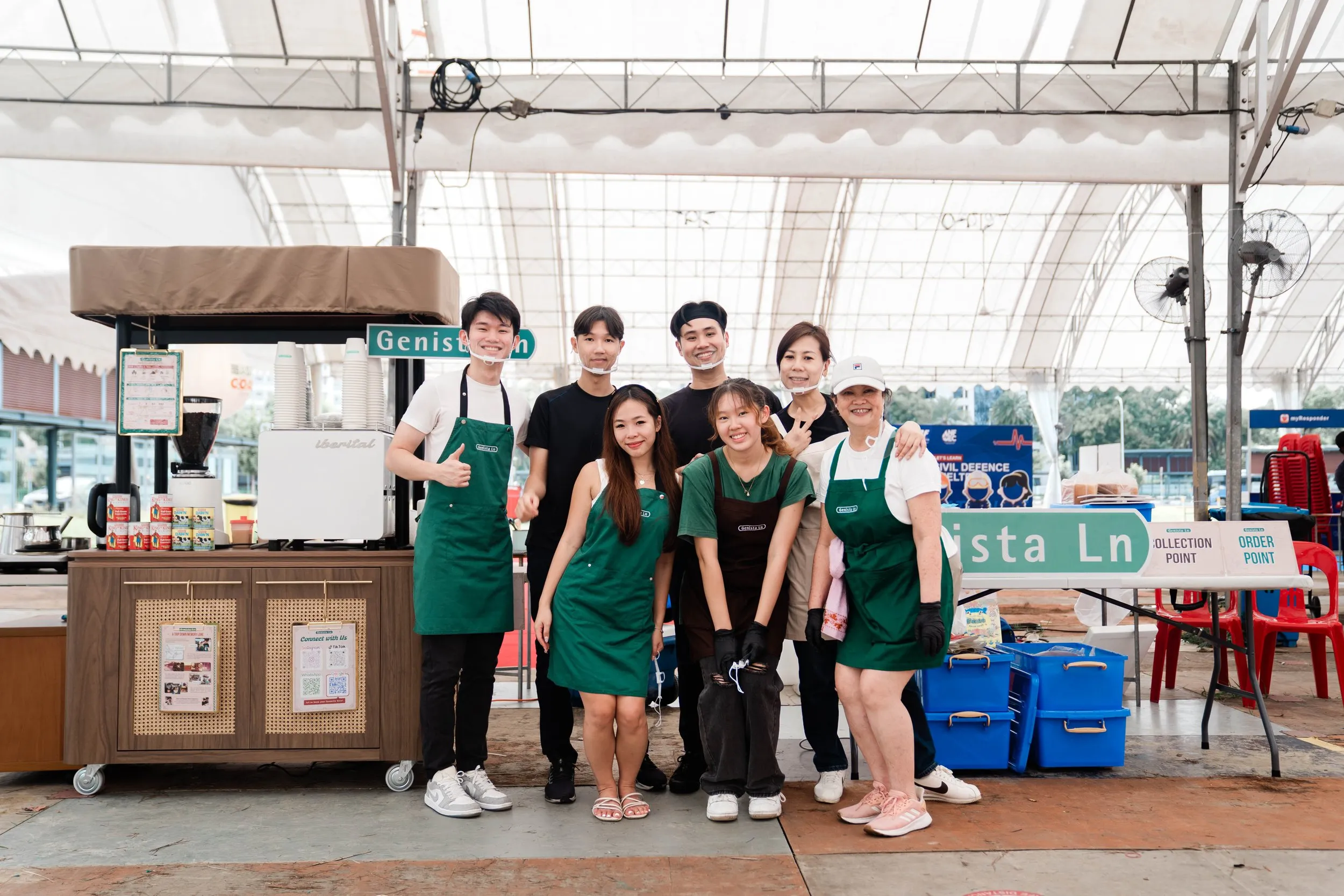 The Genista Ln crew standing together in green aprons in front of the cart under a large event marquee, with Order Point and Collection Point signs on the table beside them.