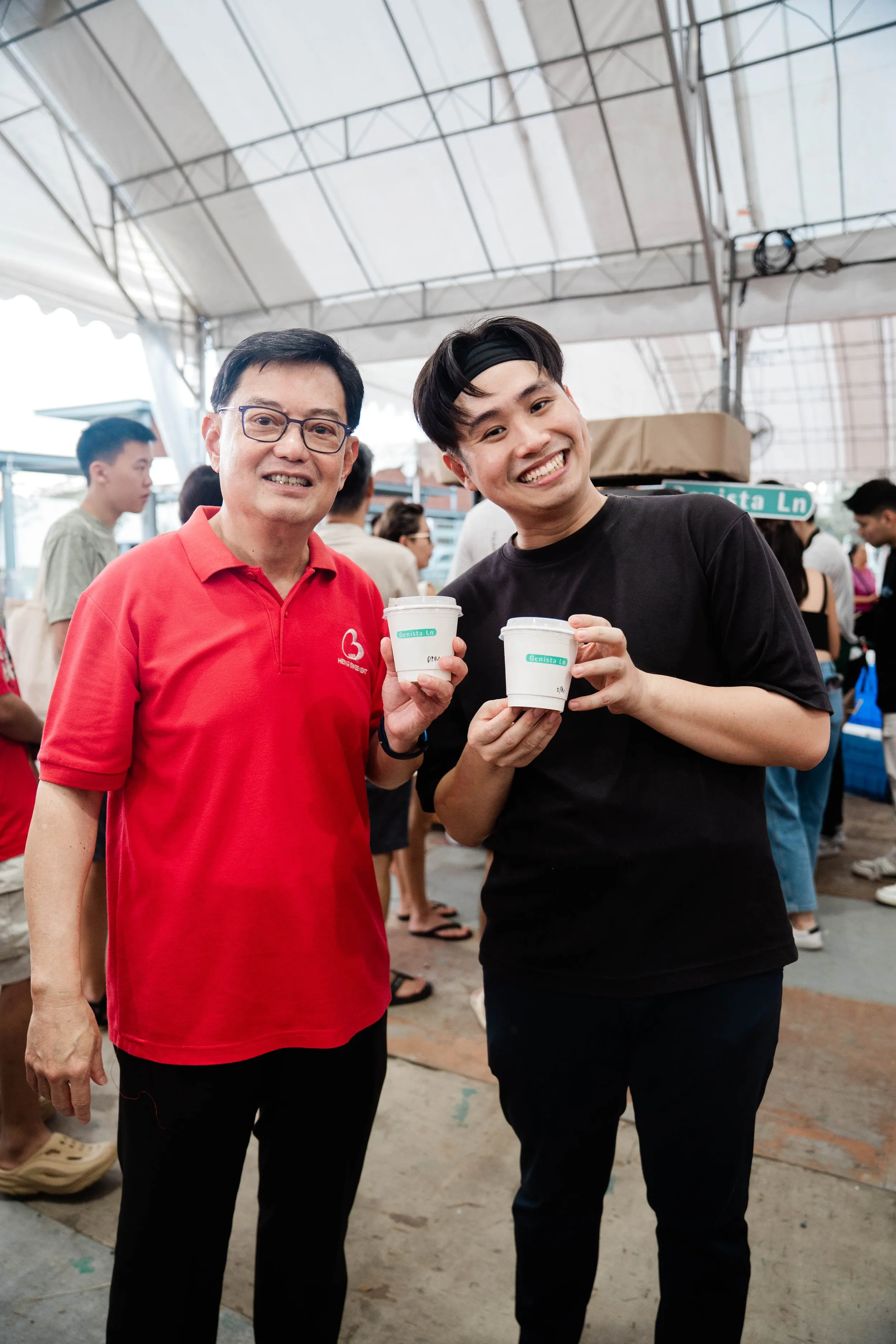 Two men smiling and holding cups of coffee at a busy outdoor gathering under a tent