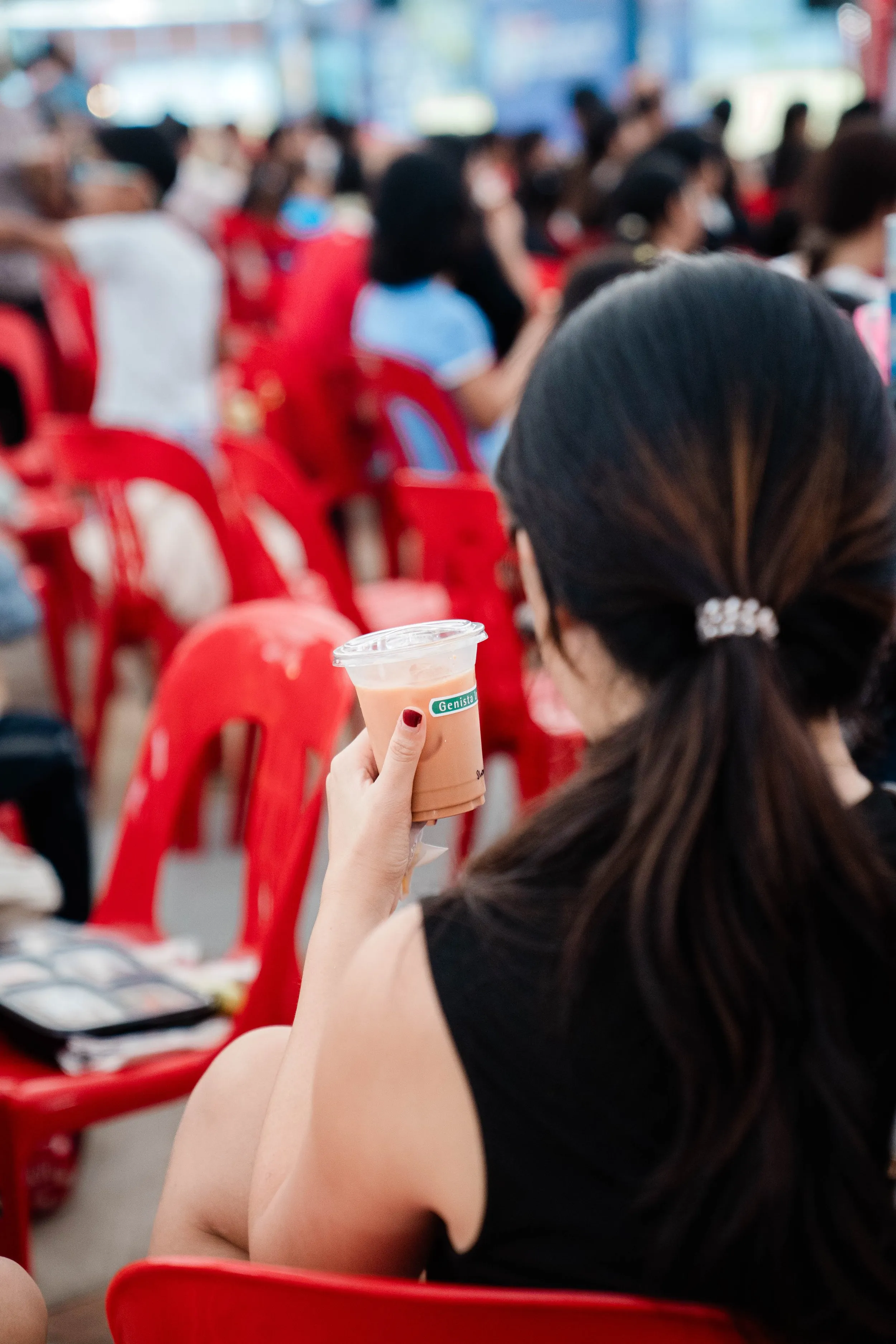 A woman with long dark hair tied back with a hair clip, wearing a black sleeveless top, is sitting on a red plastic chair in a crowded outdoor setting. She holds a cup of iced beverage with a straw, and several people are sitting on similar chairs behind her