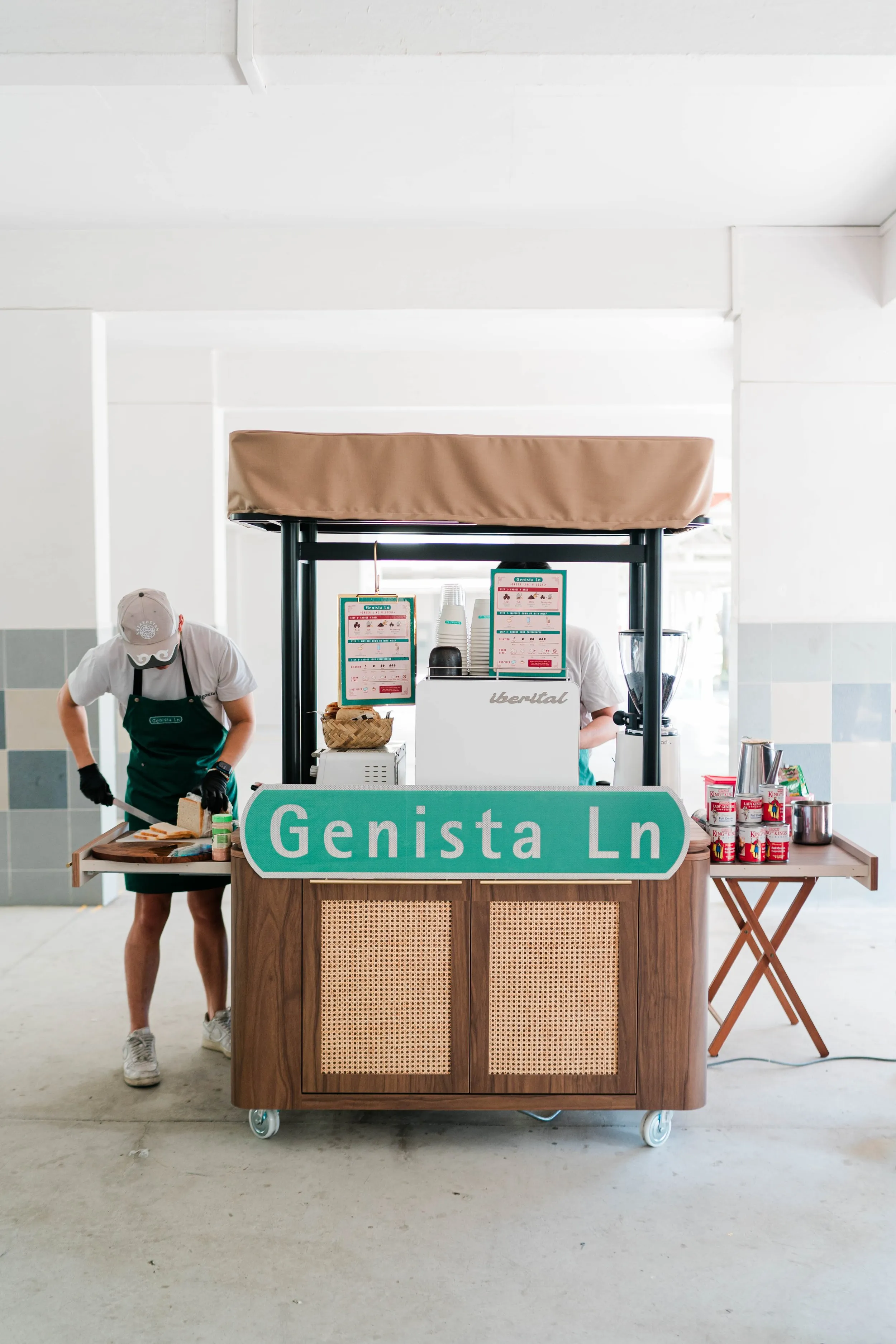 Kopi cart with the sign 'Genista Ln', two workers preparing sandwiches, cups, and canned drinks on the cart