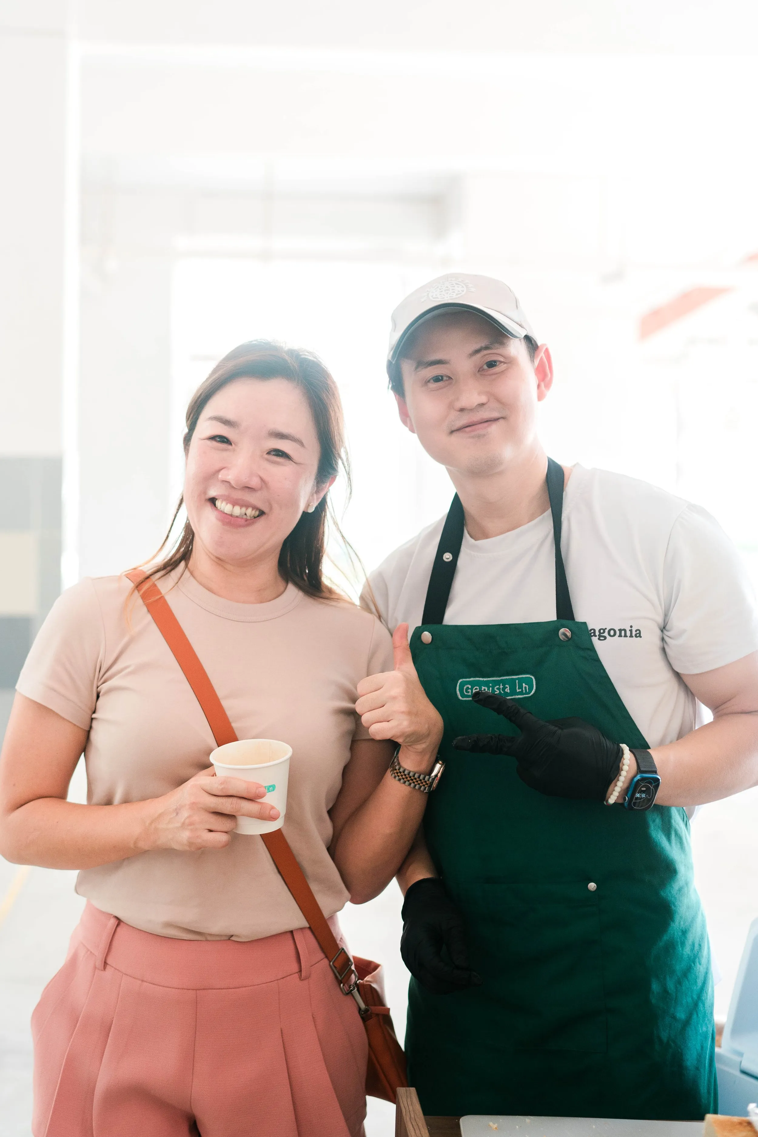 A smiling woman holding a paper cup standing next to a man in a green apron making a peace sign in a bright indoor setting