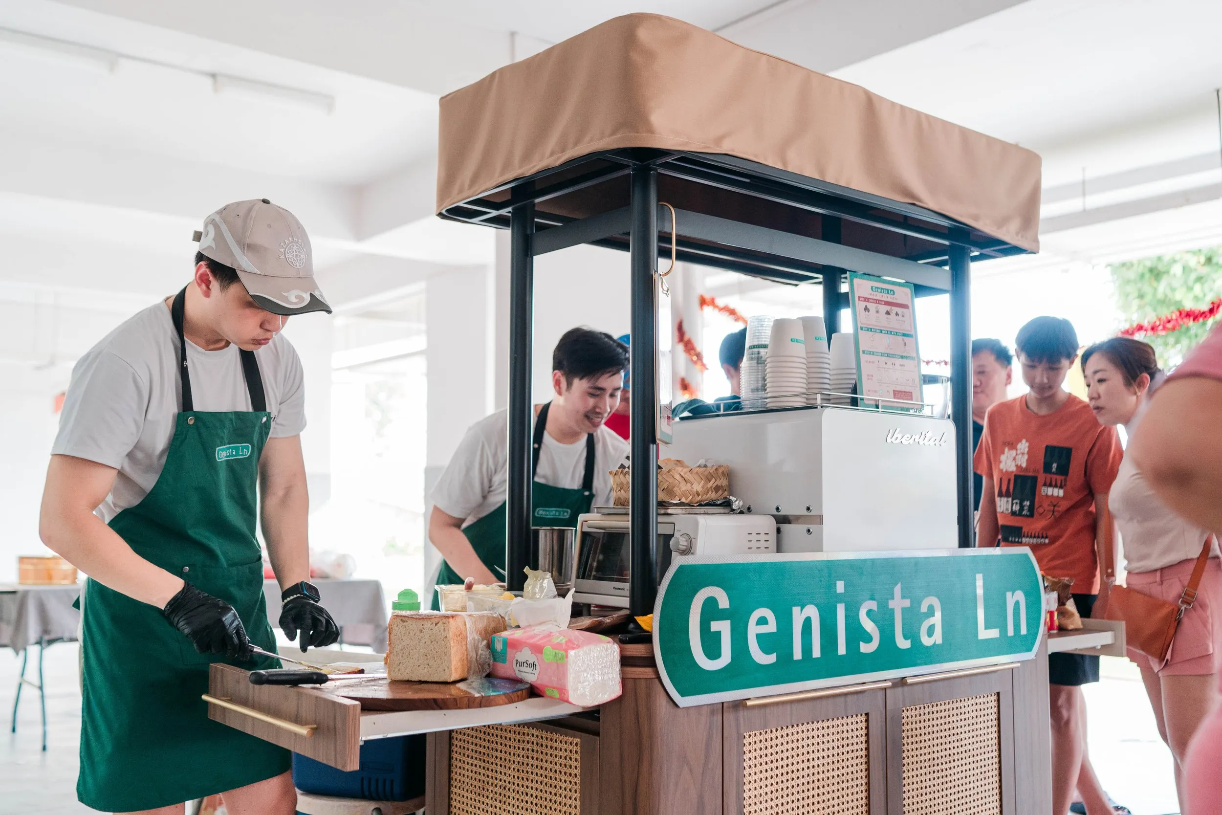 People preparing food at a street food stall with a sign that reads 'Genista Ln' in an indoor setting