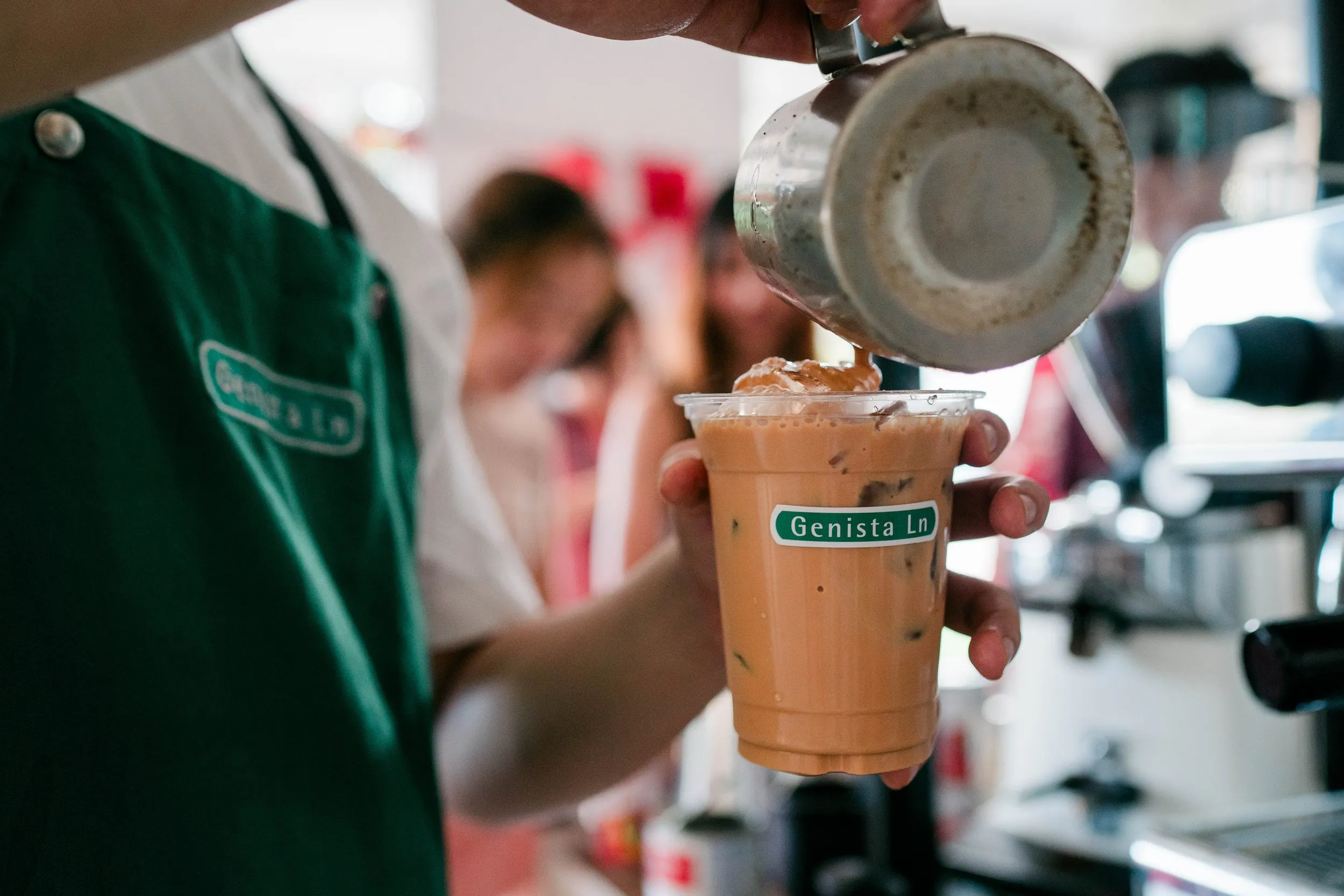 Barista pouring ice into a plastic cup of iced coffee or tea at a cafe, with other customers in the background