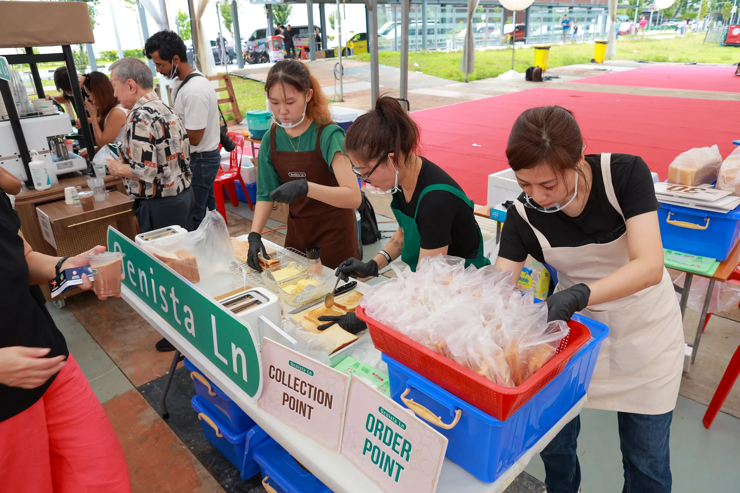 Three women working at a food stall with signs labeled 'Collection Point' and 'Order Point', serving customers at an event, with people in the background