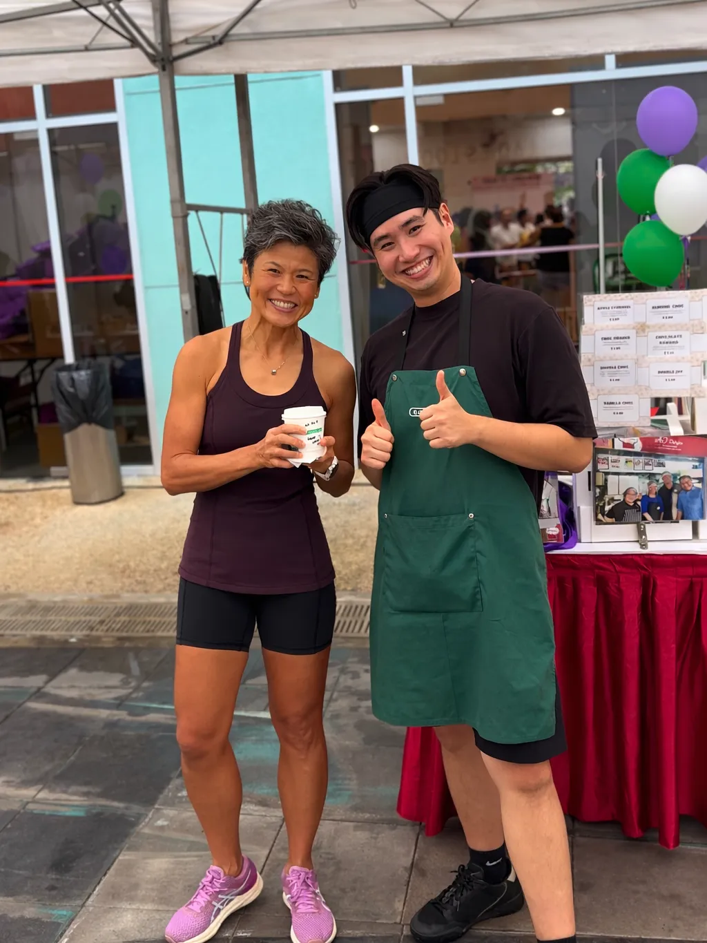 A guest in sportswear holding a Genista Ln cup beside a crew member in a green apron, under a canopy outside the community club with balloons and a skirted serving table behind them