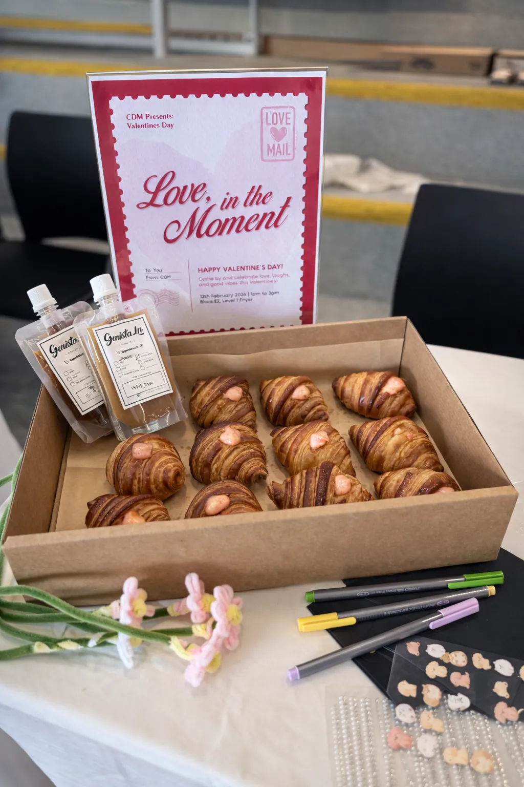 A tray of pastries and two Genista Ln kopi pouches on a table beside a Valentine's Day poster, with marker pens and paper flowers laid out alongside